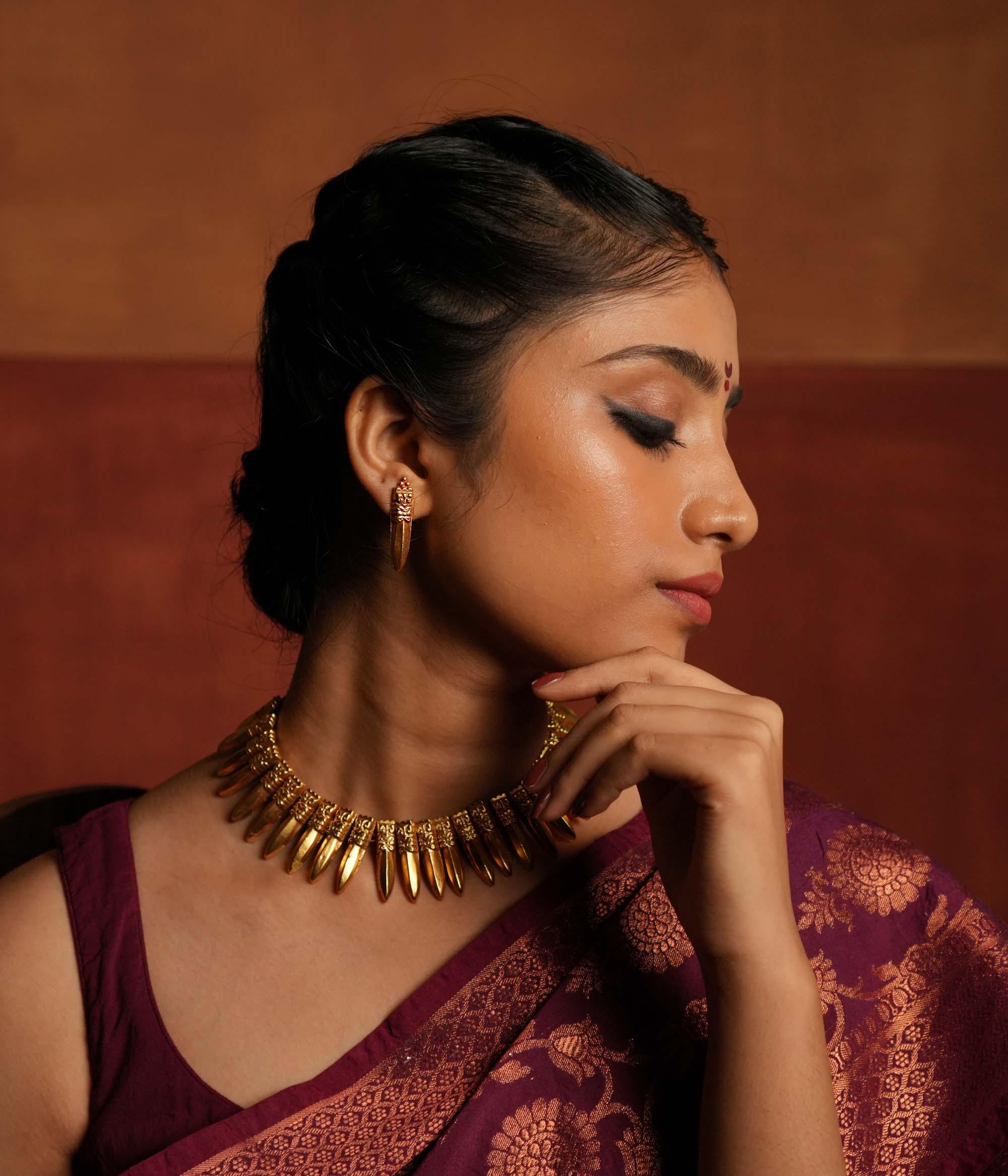 A close-up of a gold-plated earring inspired by traditional Indian designs, featuring an intricately carved top with a fierce face motif and a sleek, pointed pendant drop, worn with an elegant updo hairstyle