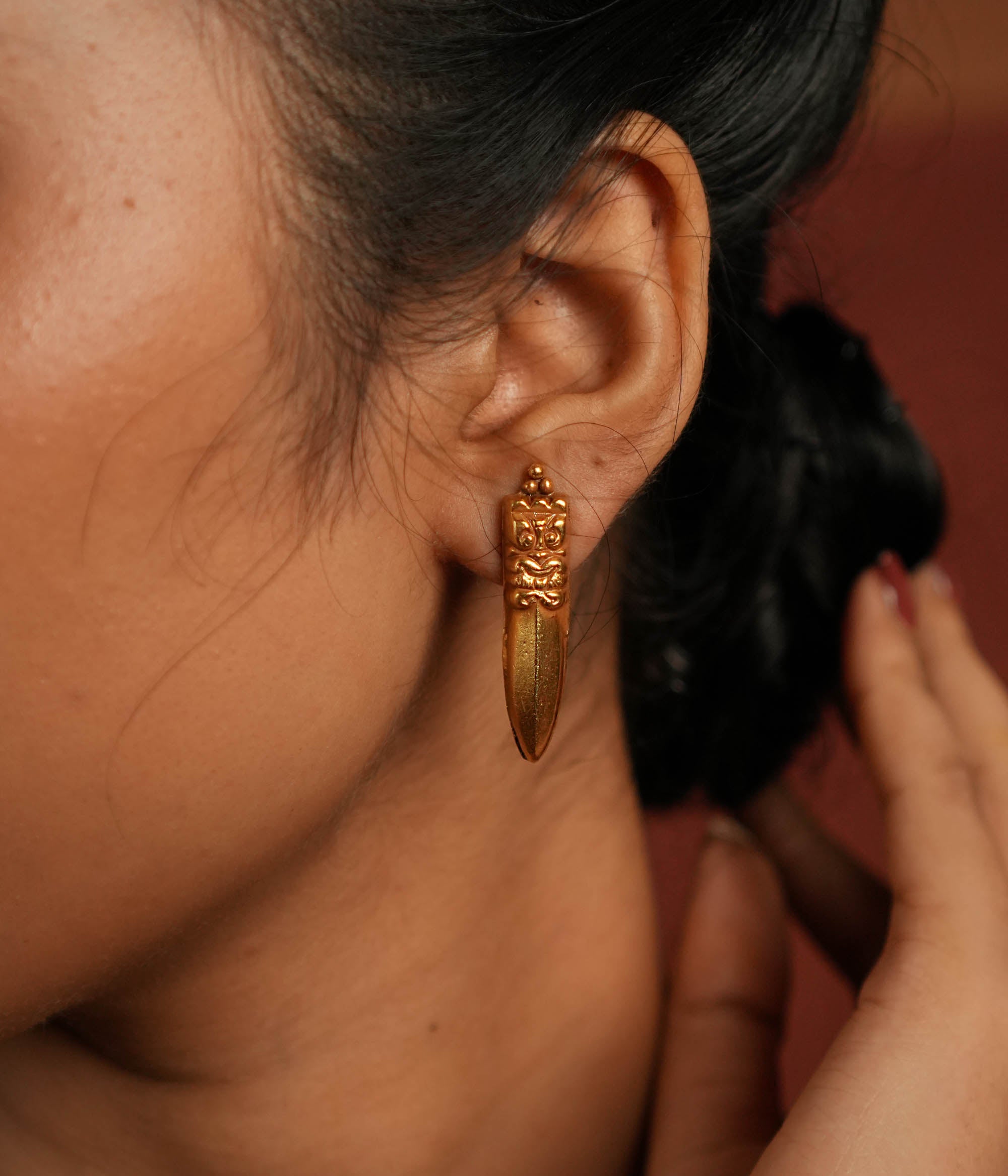 A close-up of a gold-plated earring inspired by traditional Indian designs, featuring an intricately carved top with a fierce face motif and a sleek, pointed pendant drop, worn with an elegant updo hairstyle
