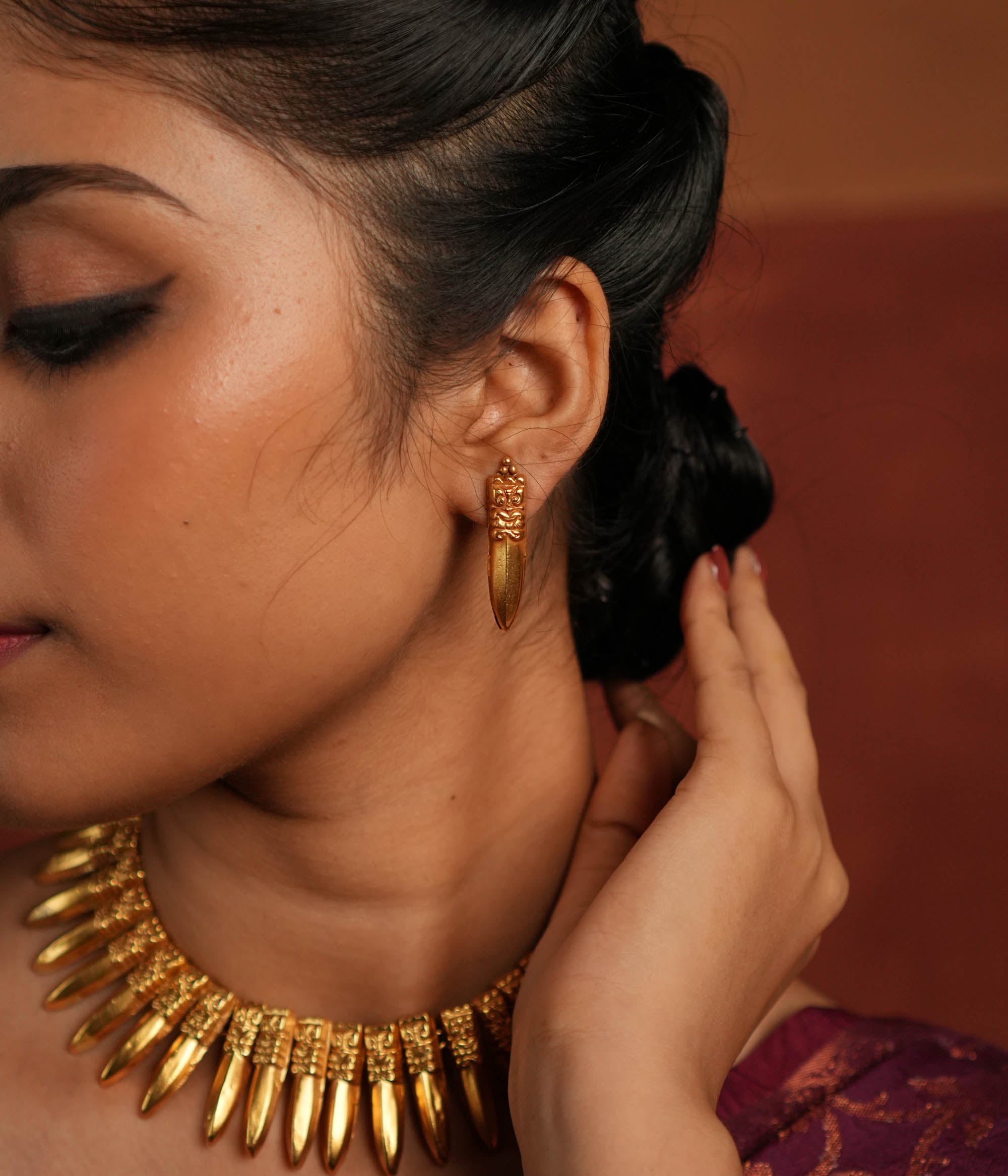 A close-up of a gold-plated earring inspired by traditional Indian designs, featuring an intricately carved top with a fierce face motif and a sleek, pointed pendant drop, worn with an elegant updo hairstyle