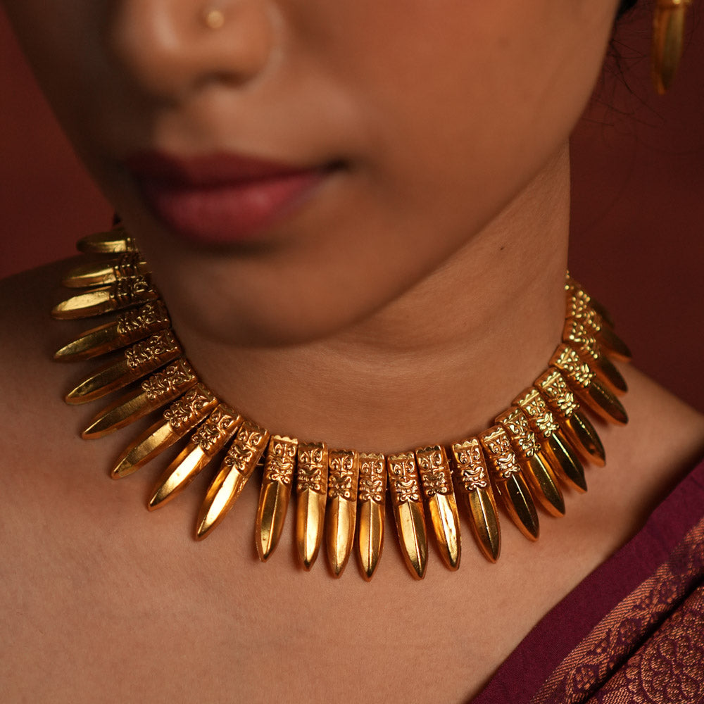 A close-up of a gold-plated choker necklace inspired by traditional Indian craftsmanship, featuring intricate woven patterns, multiple layers of polished gold beads, and subtle red gemstone accents, worn with a classic silk saree.