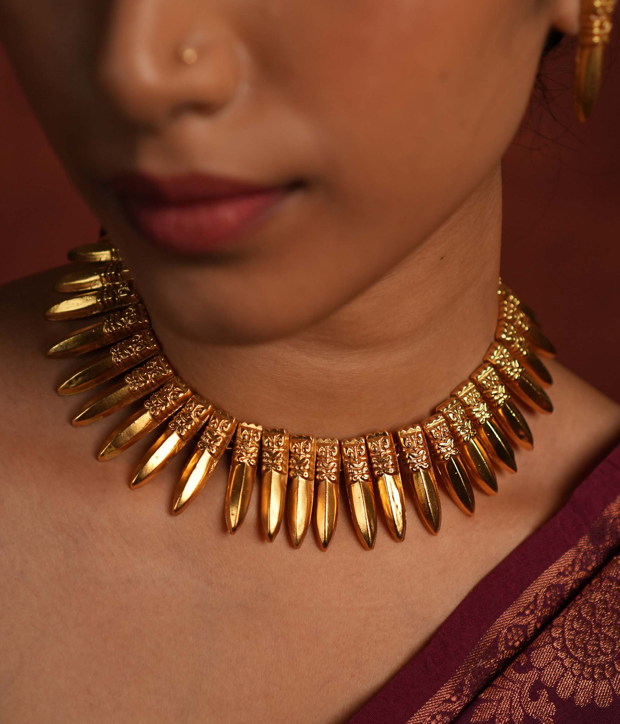 A close-up of a gold-plated choker necklace inspired by traditional Indian craftsmanship, featuring intricate woven patterns, multiple layers of polished gold beads, and subtle red gemstone accents, worn with a classic silk saree.