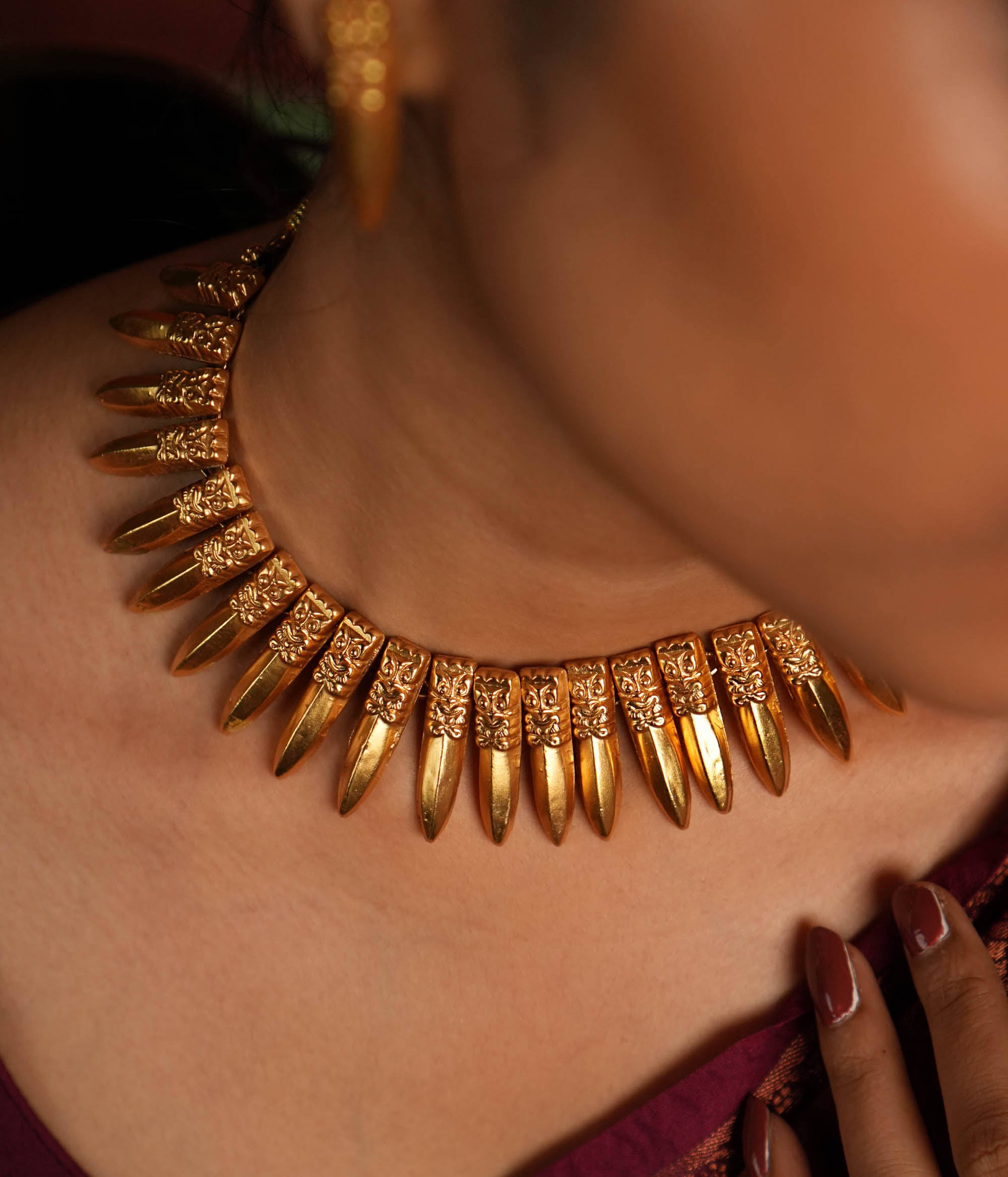 A close-up of a gold-plated choker necklace inspired by traditional Indian craftsmanship, featuring intricate woven patterns, multiple layers of polished gold beads, and subtle red gemstone accents, worn with a classic silk saree.