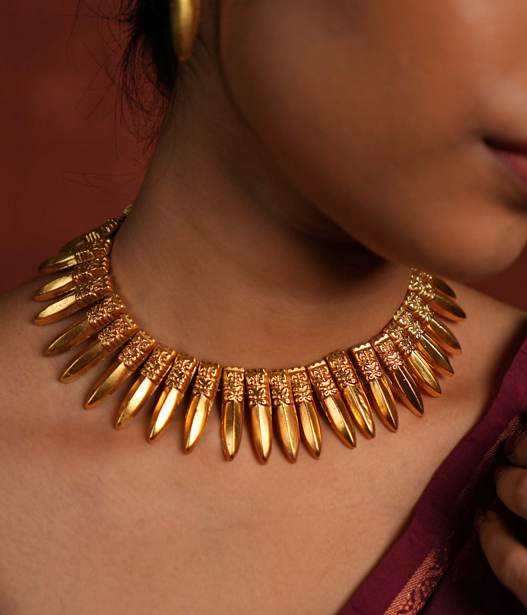 A close-up of a gold-plated choker necklace inspired by traditional Indian craftsmanship, featuring intricate woven patterns, multiple layers of polished gold beads, and subtle red gemstone accents, worn with a classic silk saree.