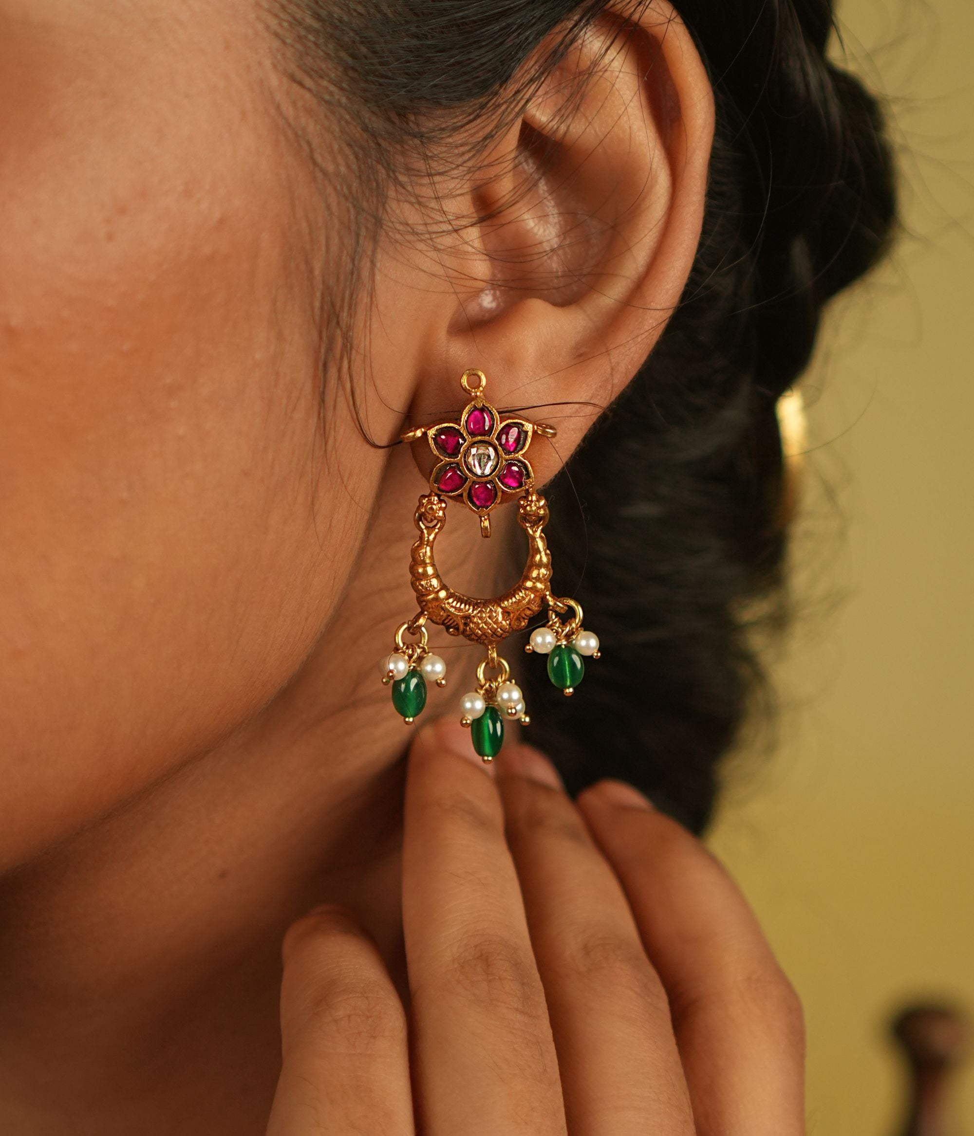 A radiant dangler earring adorned with vibrant red and green Kundan stones, where delicate petals shimmer with grace, finished with white pearl and green bead droplets for timeless allure.