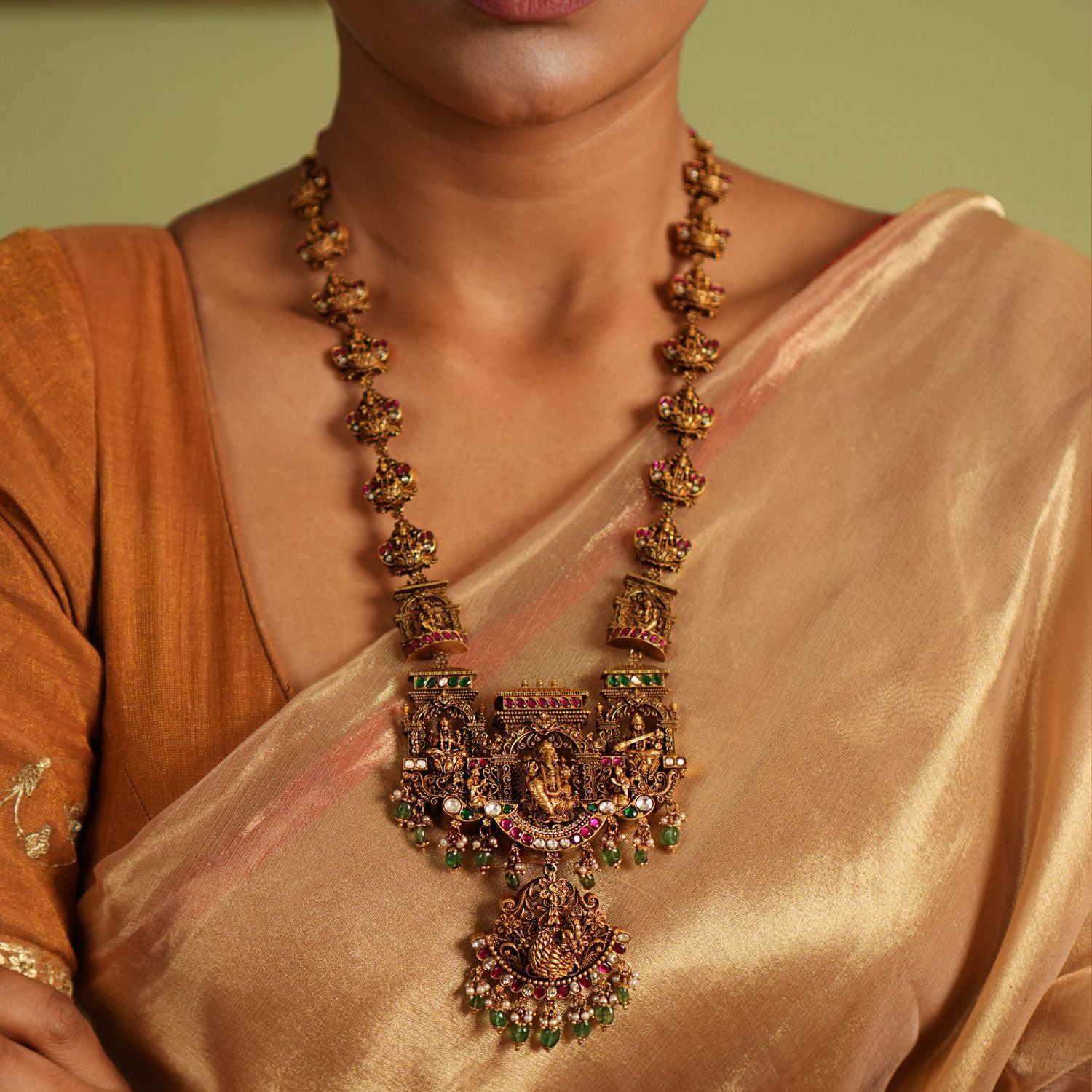 Traditional gold-plated silver temple necklace adorned with intricate carvings of deities, highlighted by Green Kundan Stones, Red Kundan Stones, pearls, and green beads. The statement centerpiece features Lord Ganesh , offering a timeless and divine elegance.