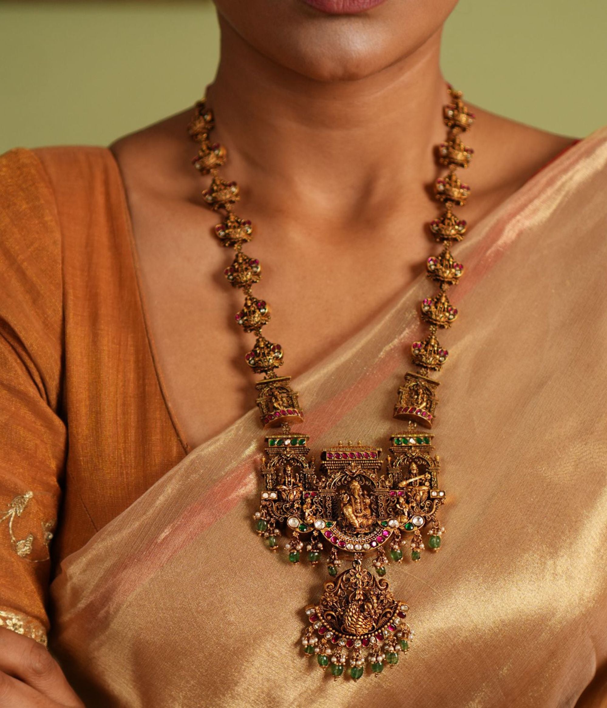 Traditional gold-plated silver temple necklace adorned with intricate carvings of deities, highlighted by Green Kundan Stones, Red Kundan Stones, pearls, and green beads. The statement centerpiece features Lord Ganesh , offering a timeless and divine elegance.