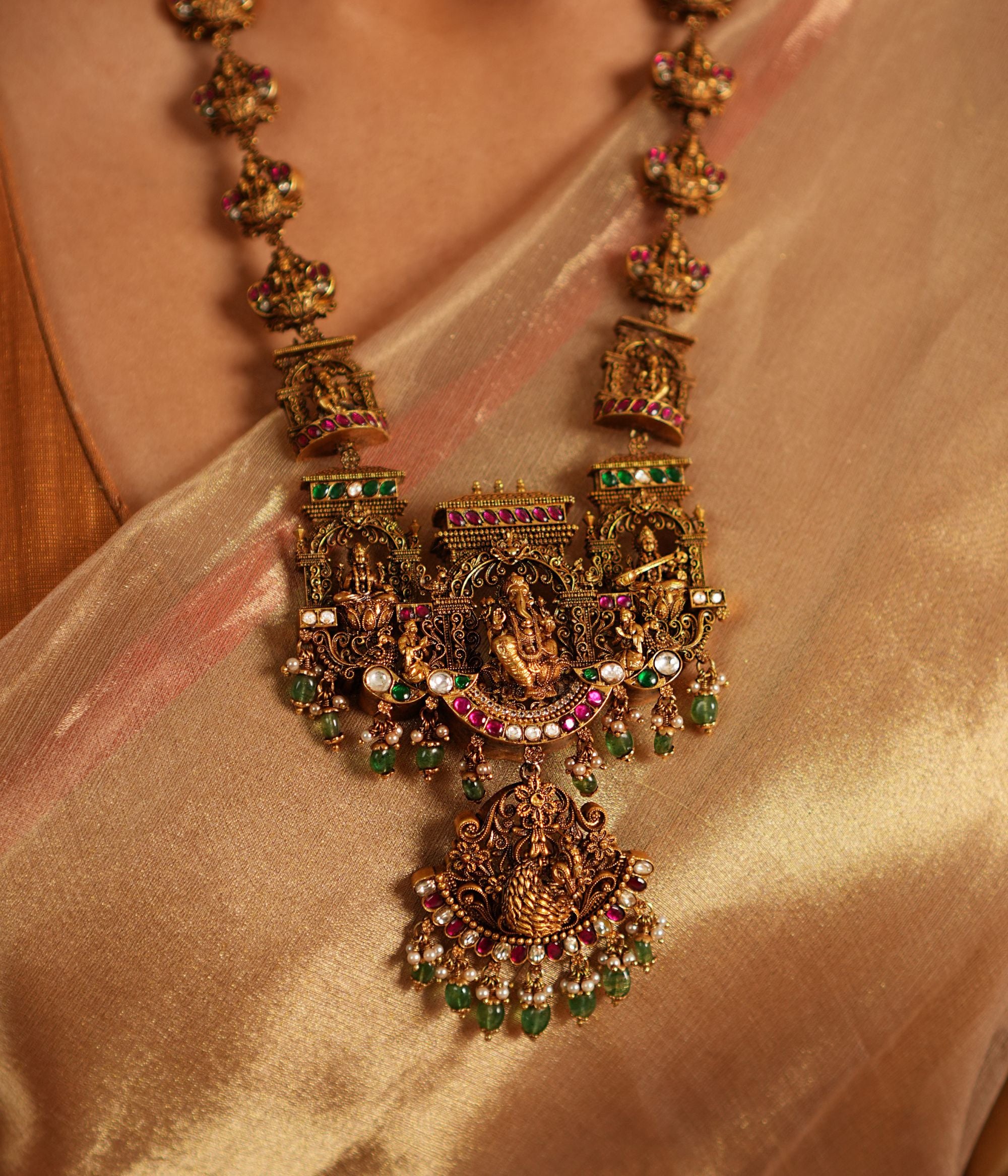 Traditional gold-plated silver temple necklace adorned with intricate carvings of deities, highlighted by Green Kundan Stones, Red Kundan Stones, pearls, and green beads. The statement centerpiece features Lord Ganesh , offering a timeless and divine elegance.