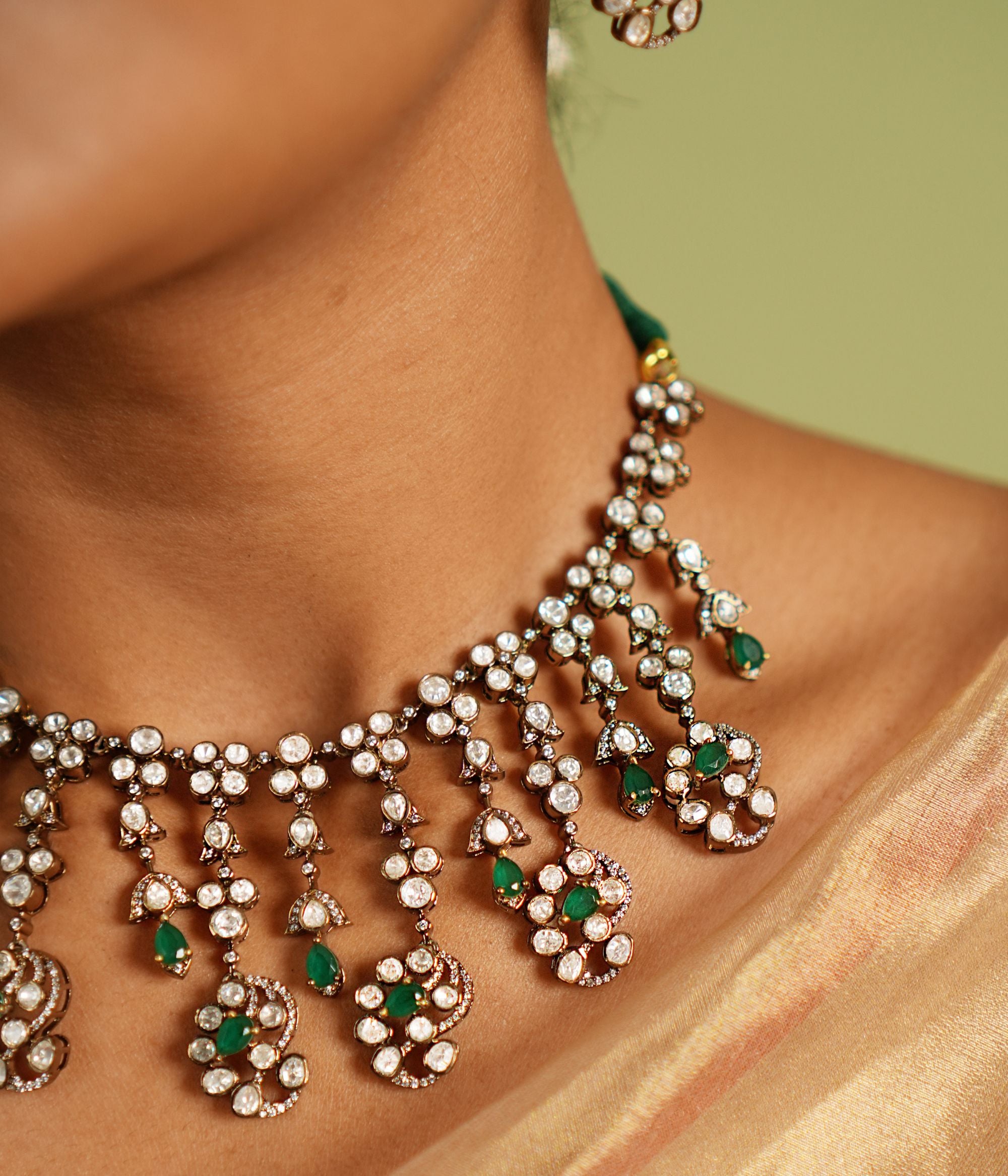 Echoes of a bygone era reborn! This gold-plated silver necklace features floral-shaped droplets adorned with Polki, CZ stones, and green spinal gems, cascading like delicate vines. A timeless blend of Victorian charm and graceful allure.