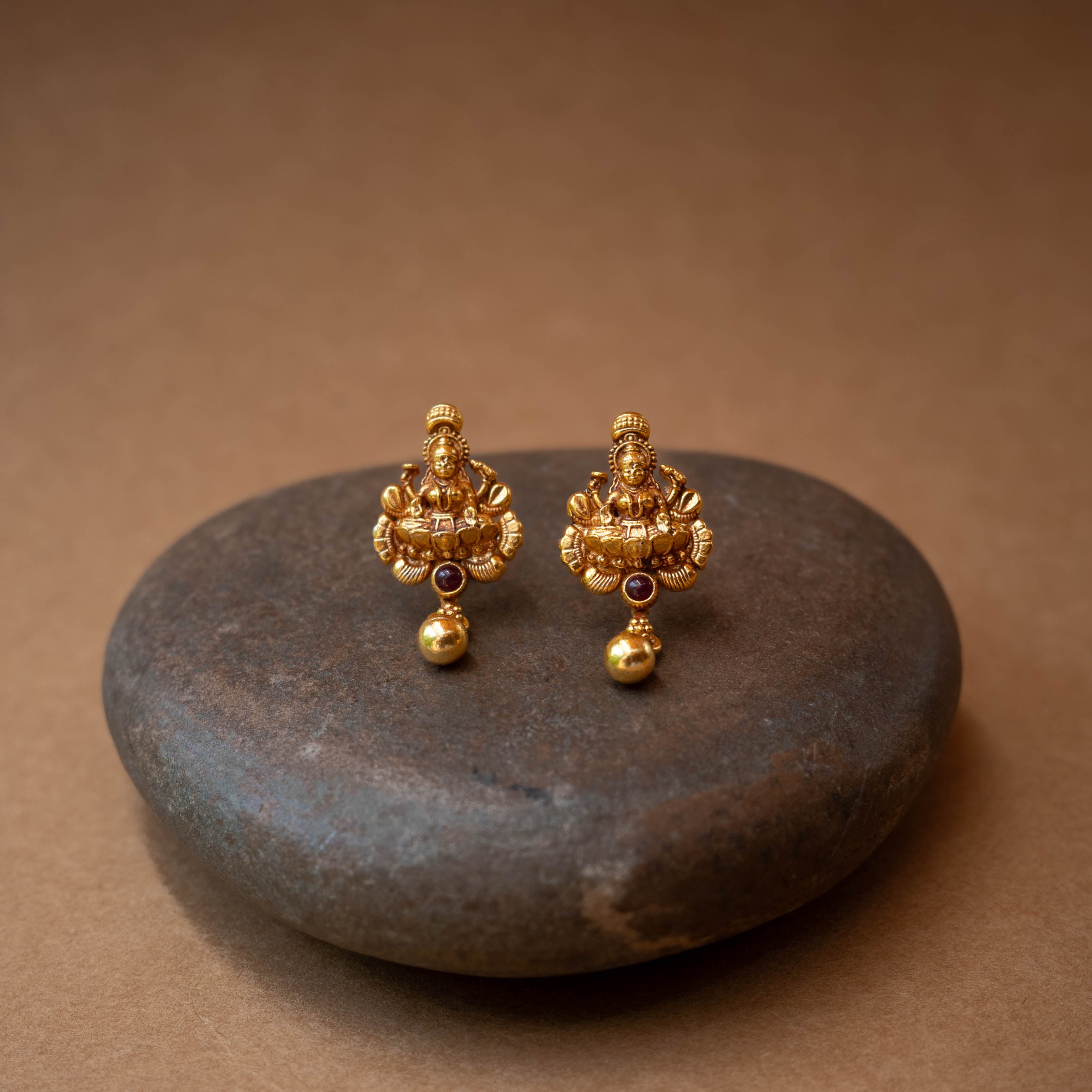 Traditional stud earrings with a goddess figure, a red stone, and a dangling bead.