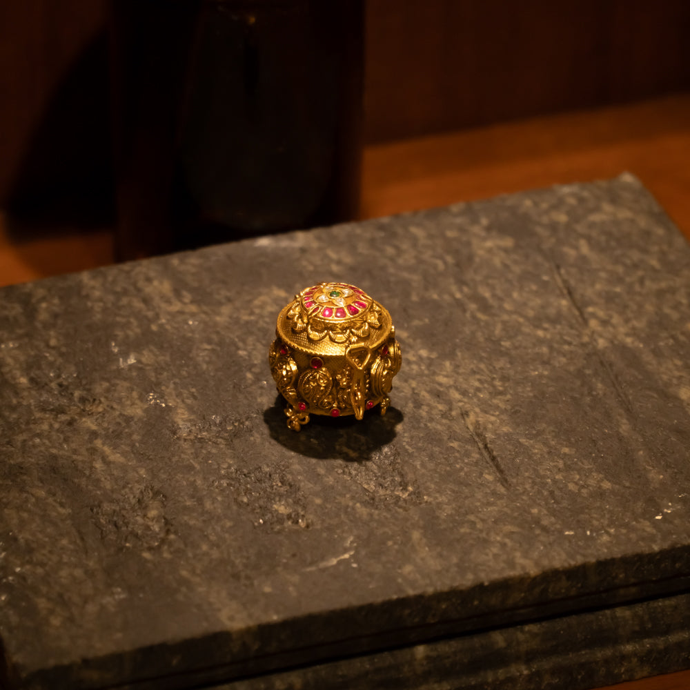 Temple-style golden kumkum box with red and green Kundan stones, placed on stone surface.