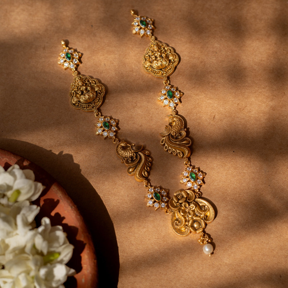 Antique gold-finished 92.5 sterling silver Peacock Devotion Antique Necklace featuring intricate Goddess Lakshmi and peacock motifs, adorned with shimmering green stones and delicate white crystals, reflecting traditional South Indian temple jewelry craftsmanship for festive occasions and weddings.