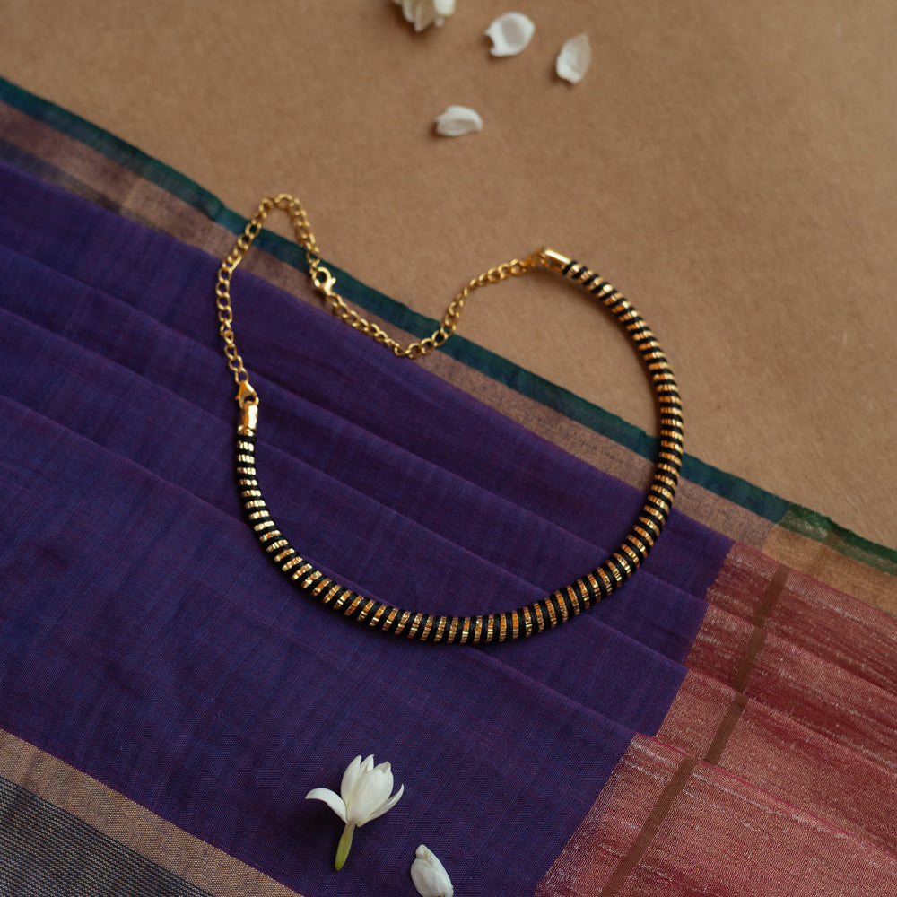 Close-up of a necklace featuring dark Rudraksha beads interspersed with thin gold accents. The necklace has a gold chain and clasp, resting on a purple and maroon fabric.