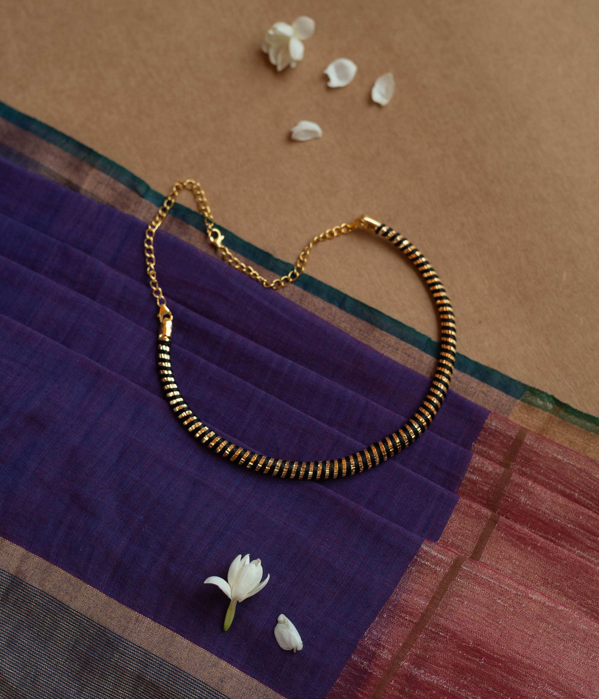 Close-up of a necklace featuring dark Rudraksha beads interspersed with thin gold accents. The necklace has a gold chain and clasp, resting on a purple and maroon fabric.