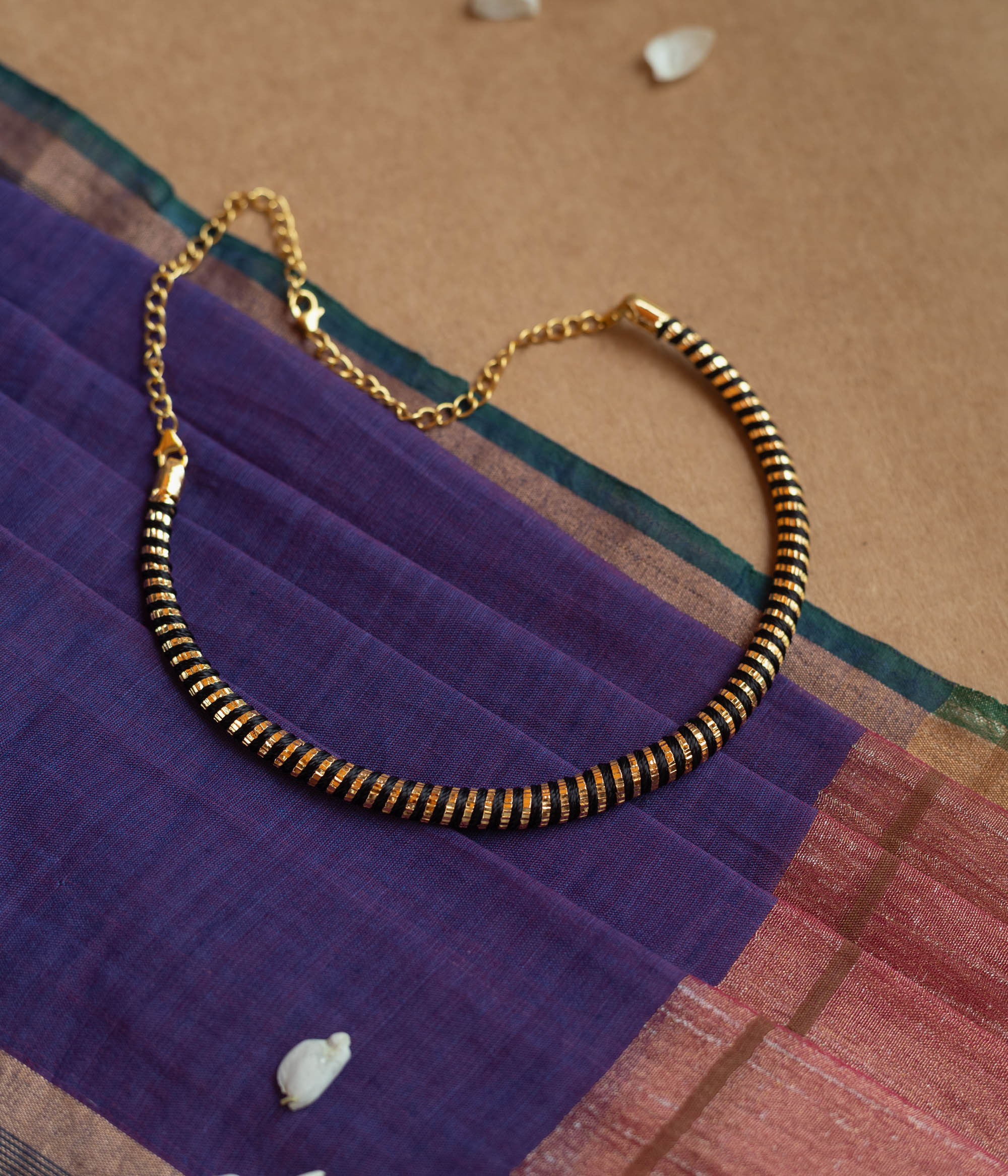Close-up of a necklace featuring dark Rudraksha beads interspersed with thin gold accents. The necklace has a gold chain and clasp, resting on a purple and maroon fabric.
