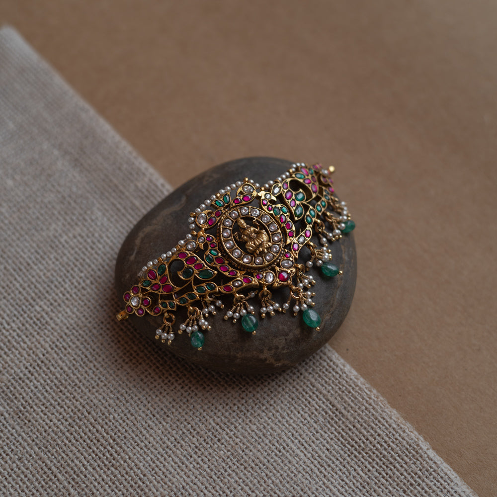 Temple-style choker with Lakshmi motif, kundan stones, pearls, and green beads on stone surface.