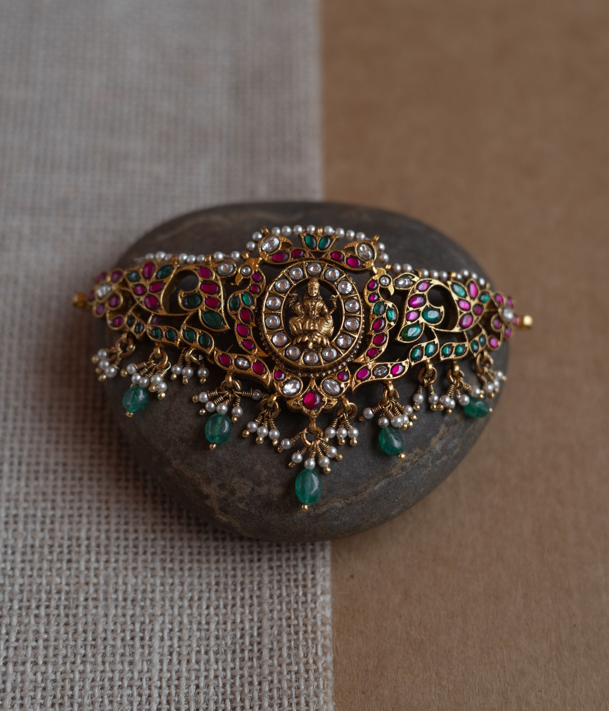 Temple-style choker with Lakshmi motif, kundan stones, pearls, and green beads on stone surface.