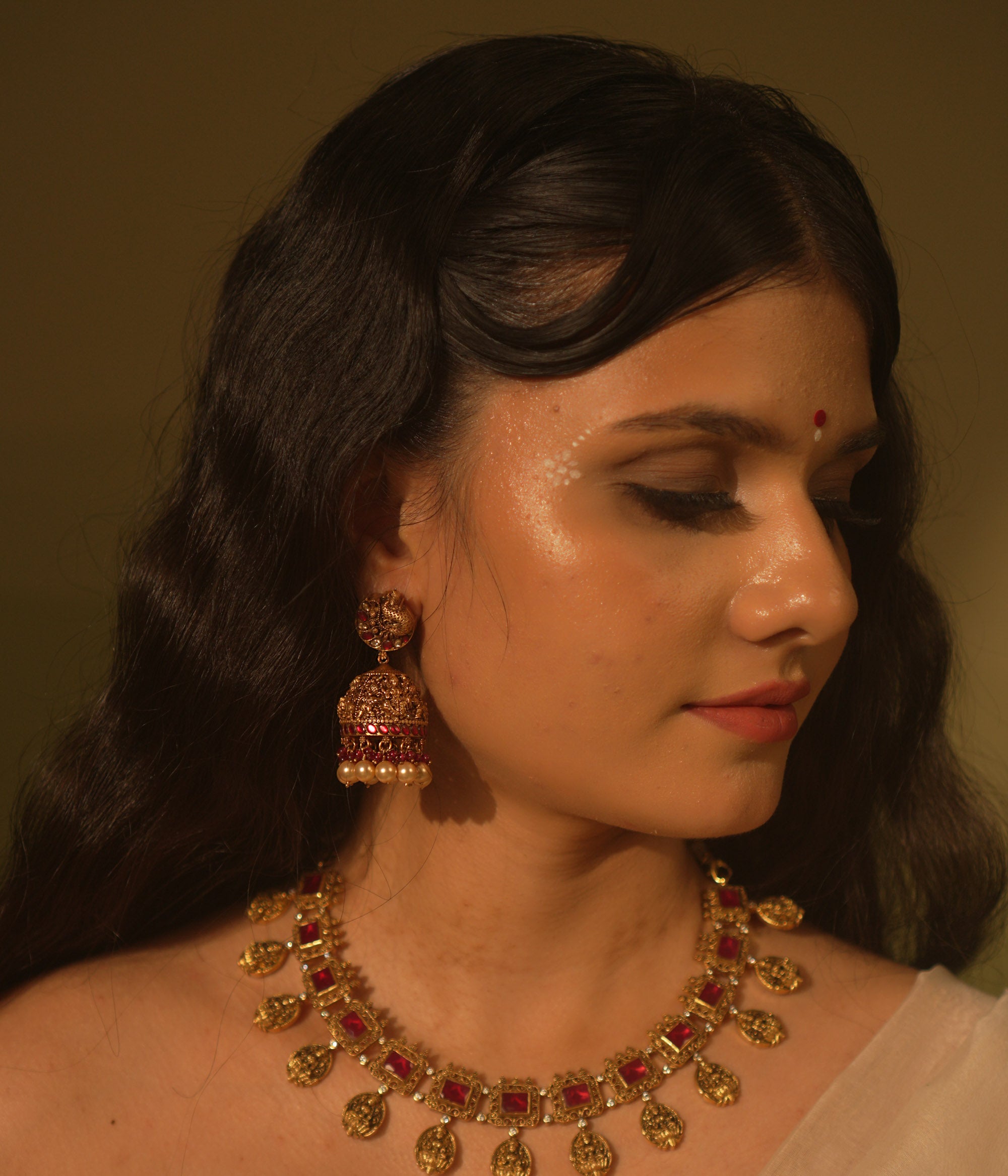 Antique gold temple jhumka earrings with peacock motif, red and green kundan, red beads, and pearl drops, on a woman's ear.