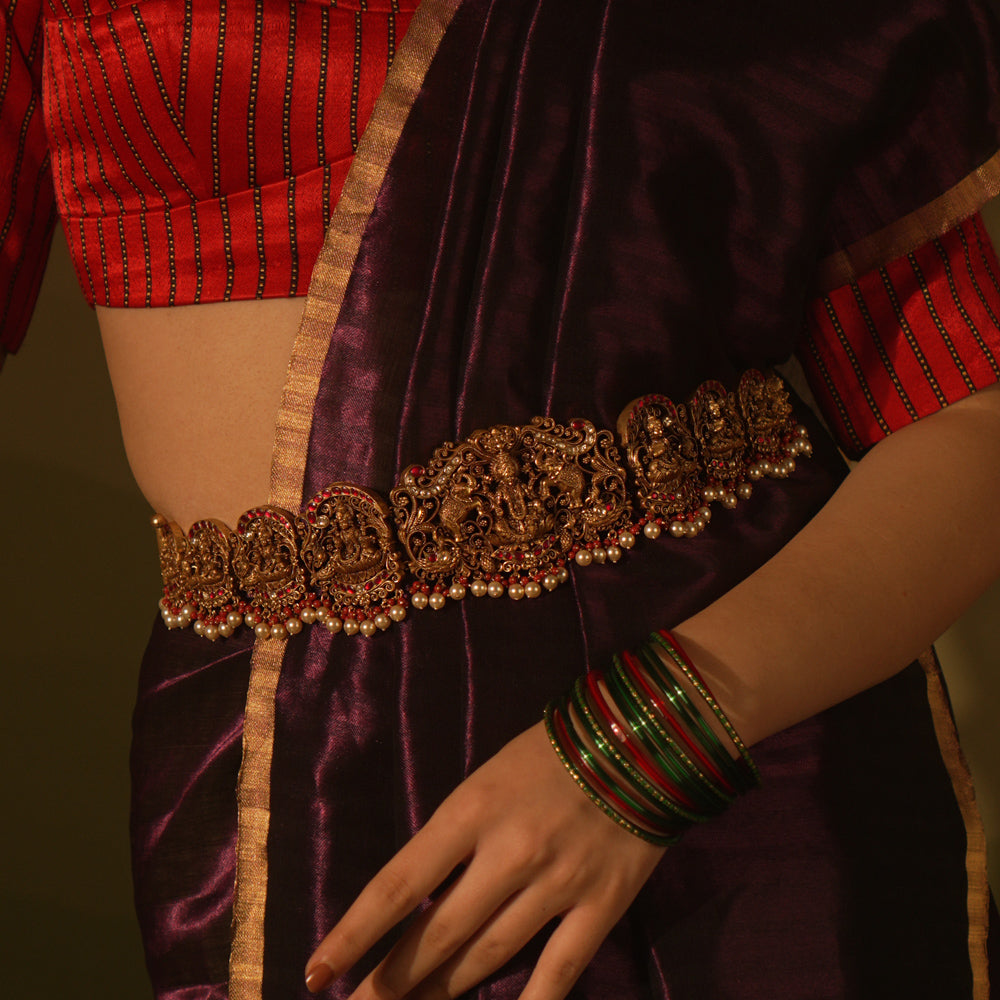 Temple vaddanam with goddess motifs, red-white Kundan stones, and pearl drops on a saree.