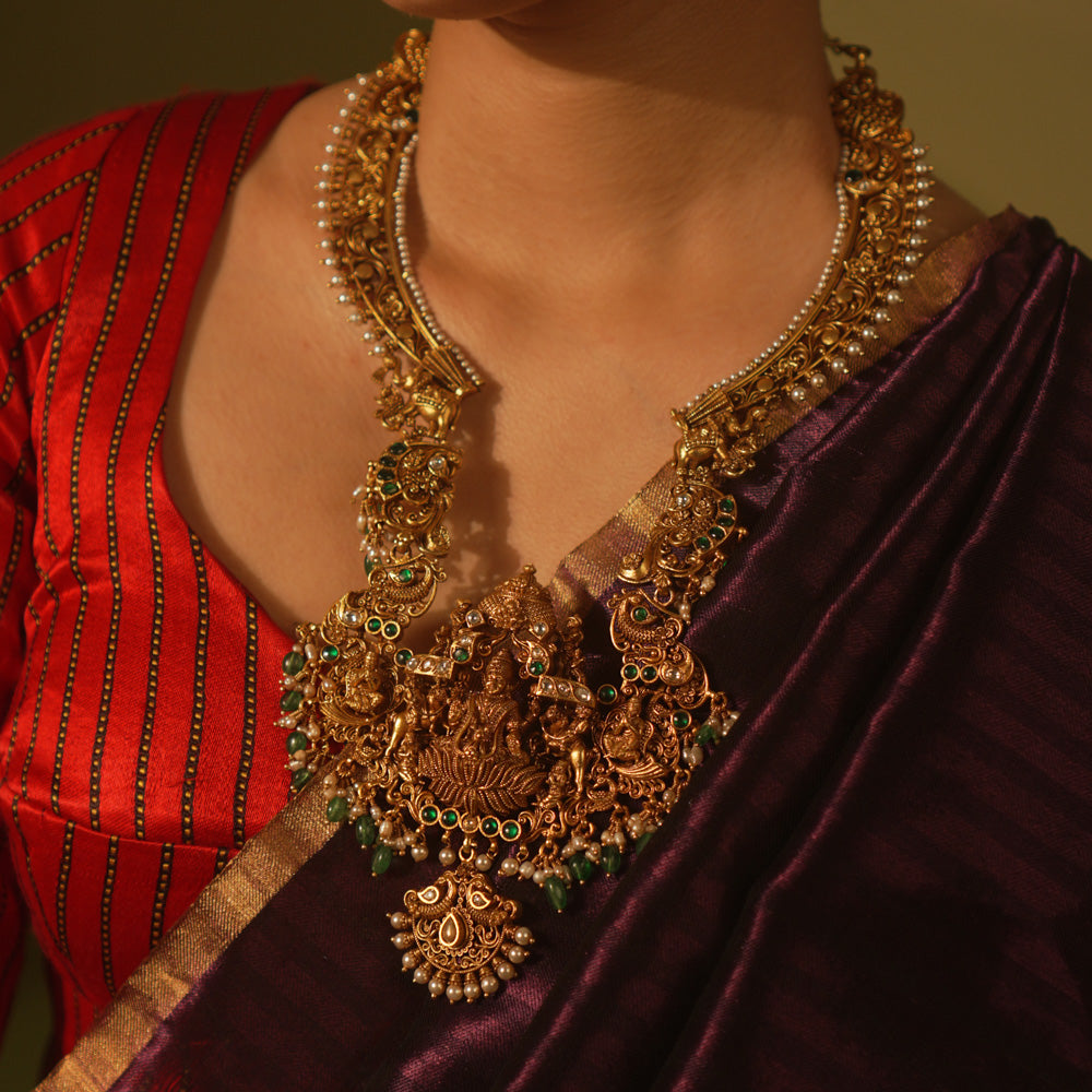 Temple-style gold necklace featuring Goddess Lakshmi, green beads, white Kundan stones, and intricate divine motifs in antique finish.