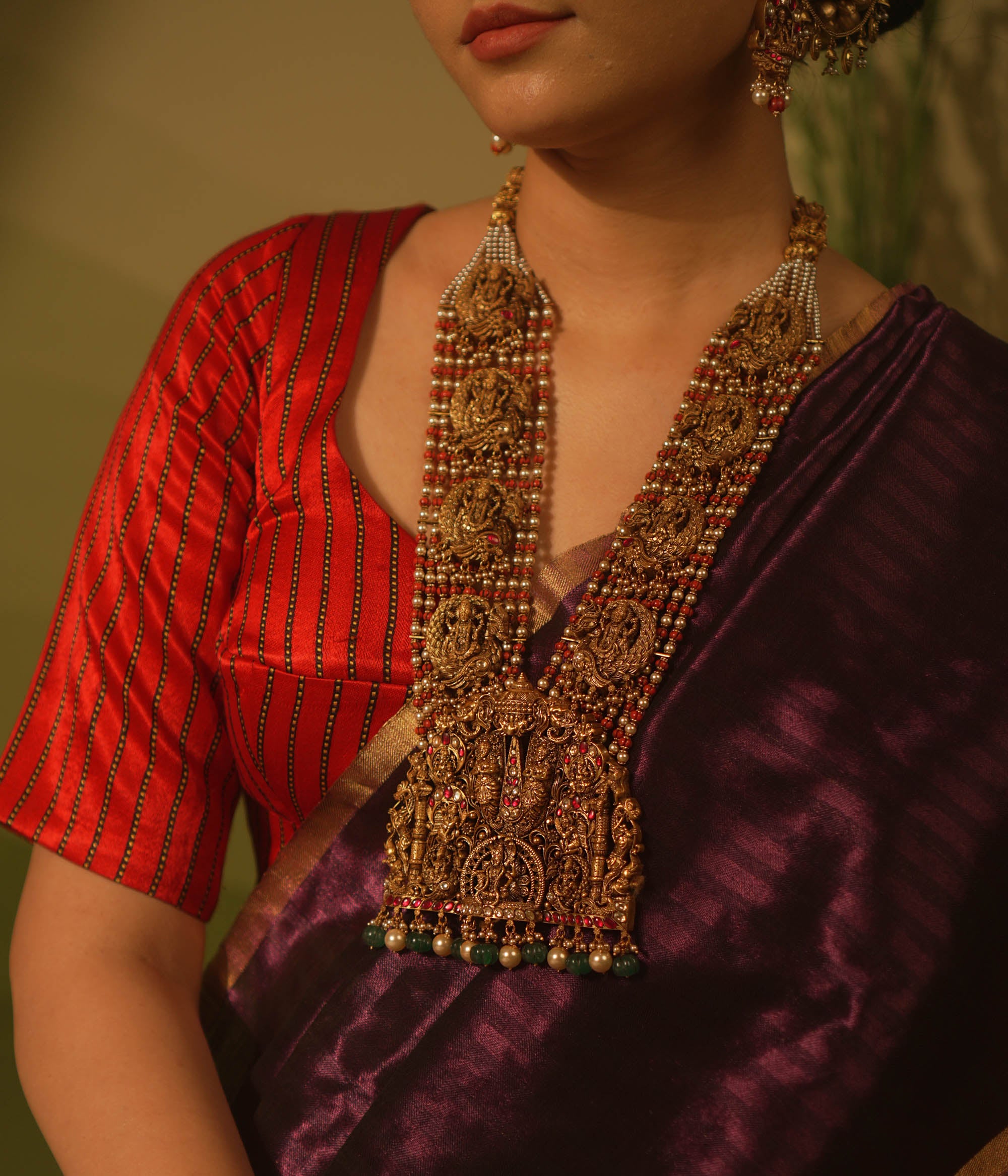 Temple necklace featuring goddess Lakshmi motifs, red and white Kundan stones, green beads, and pearls for traditional bridal wear.