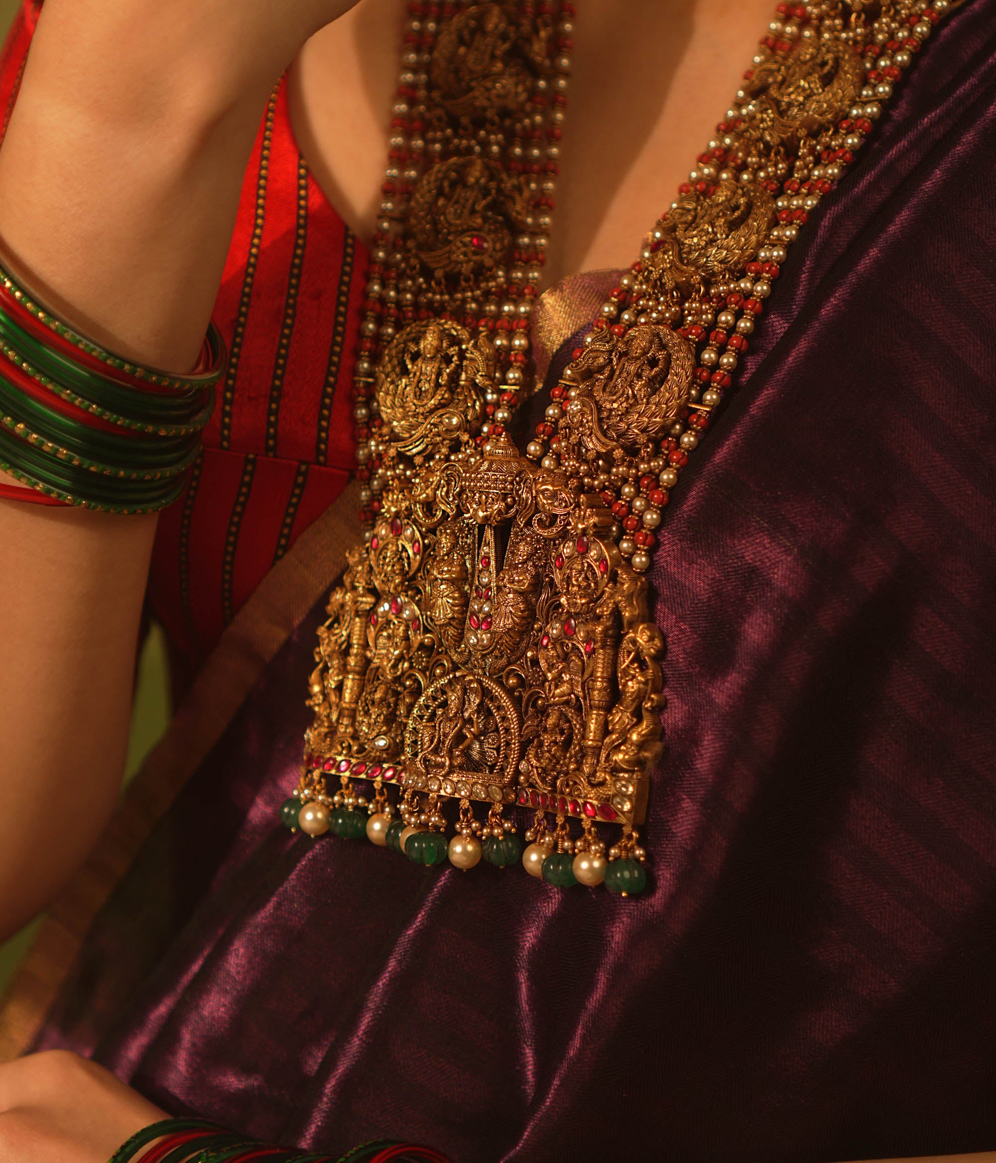 Temple necklace featuring goddess Lakshmi motifs, red and white Kundan stones, green beads, and pearls for traditional bridal wear.