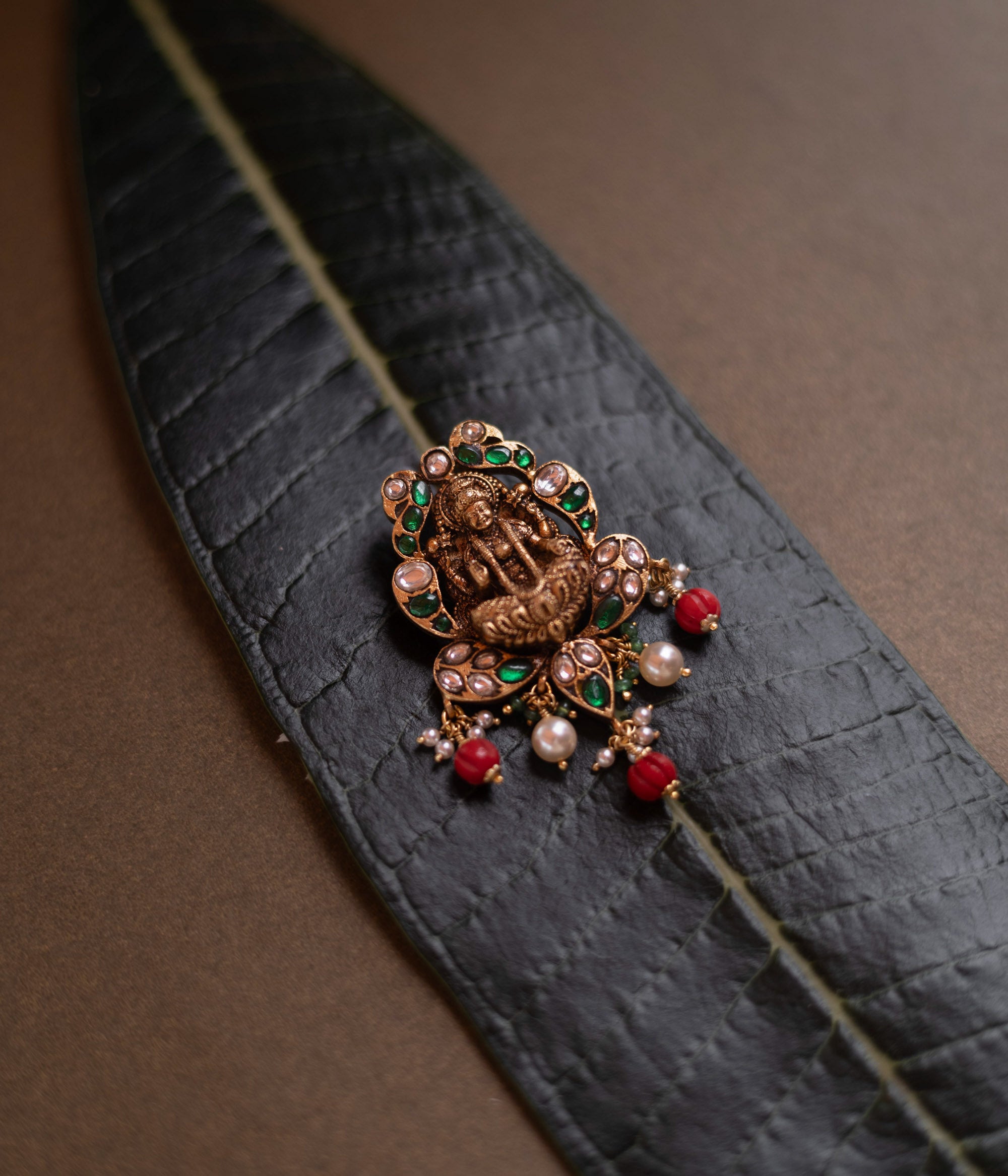 Temple-style pendant featuring Goddess Lakshmi, kundan stones, pearls, and red beads on a green leaf.