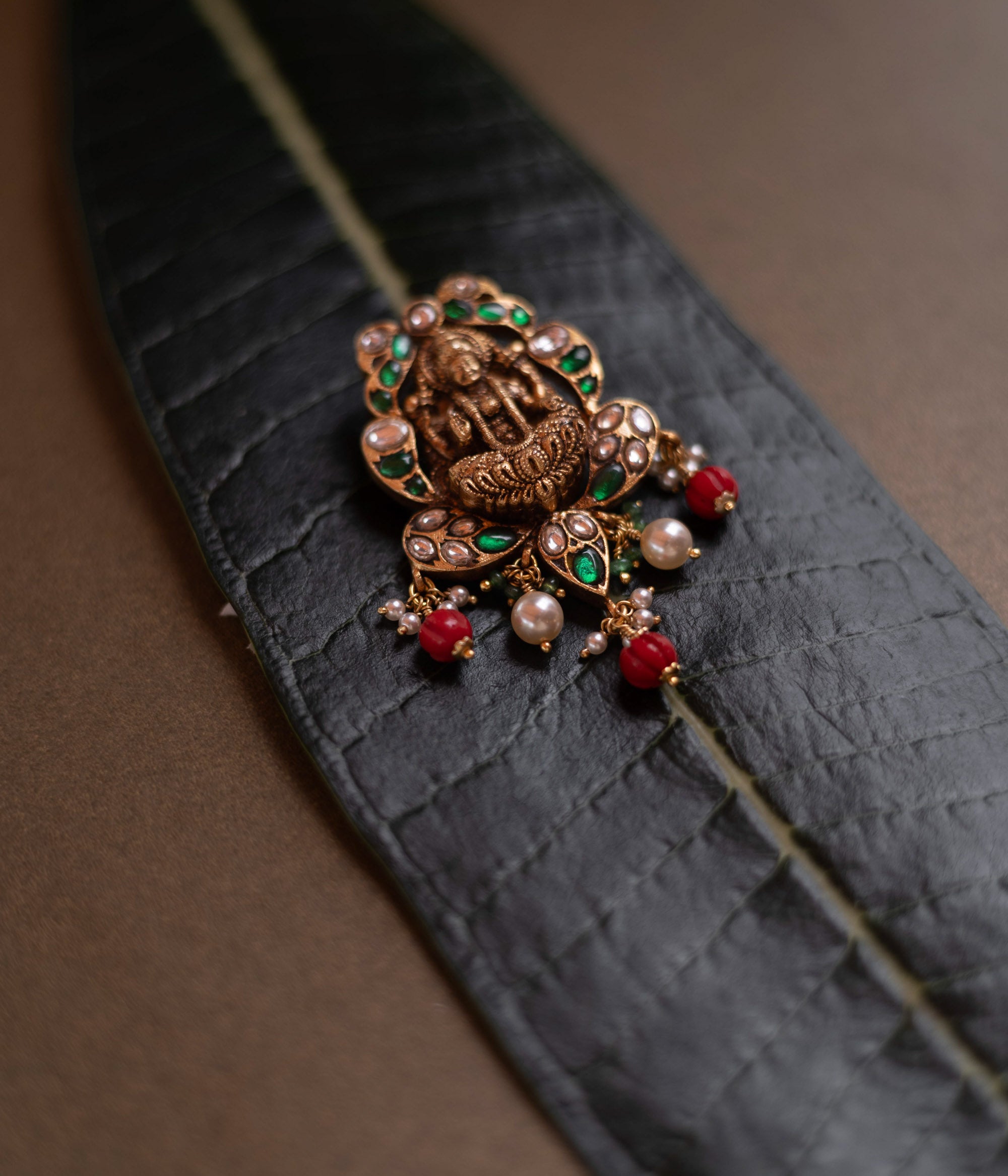 Temple-style pendant featuring Goddess Lakshmi, kundan stones, pearls, and red beads on a green leaf.