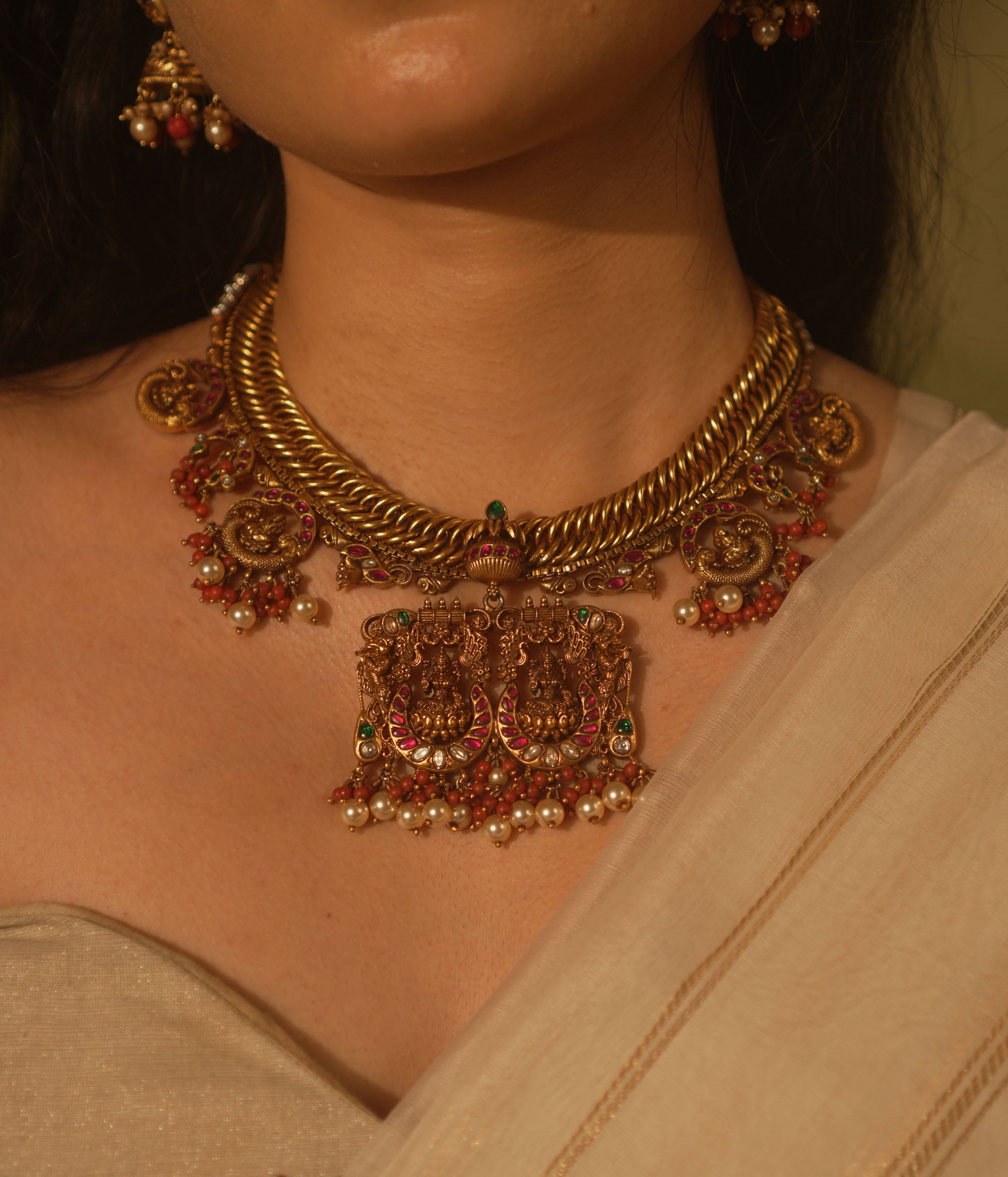 Gold temple necklace with pink kundan peacock pendants, carved deity, pearls, and coral.