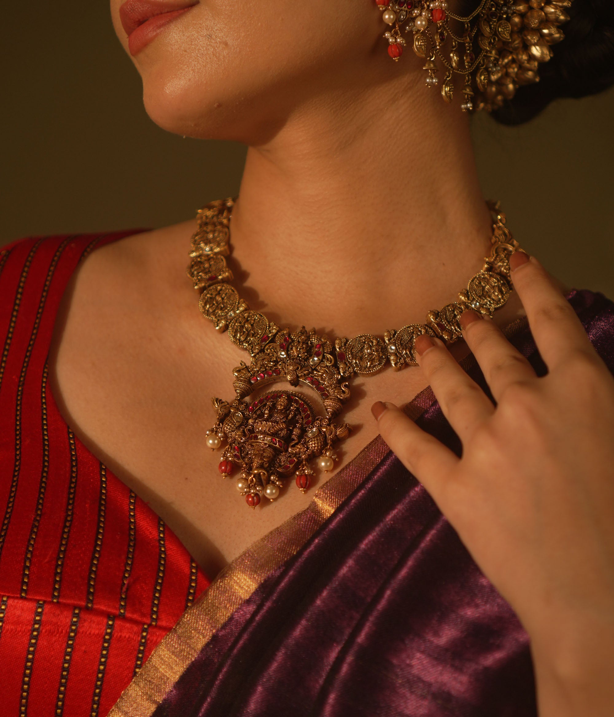 Antique gold temple necklace with goddess motifs, red and white kundan, red beads, and pearl drops, worn by a woman.