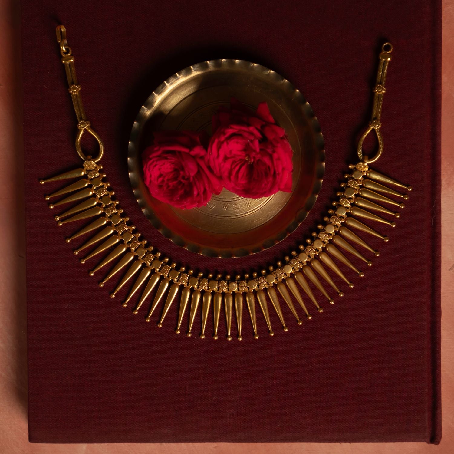 Gold Plated Silver Necklace with textured, spike-like pendants arranged in a semi-circle on a deep red surface. A brass dish and two red roses in the background enhance the rich, elegant aesthetic.