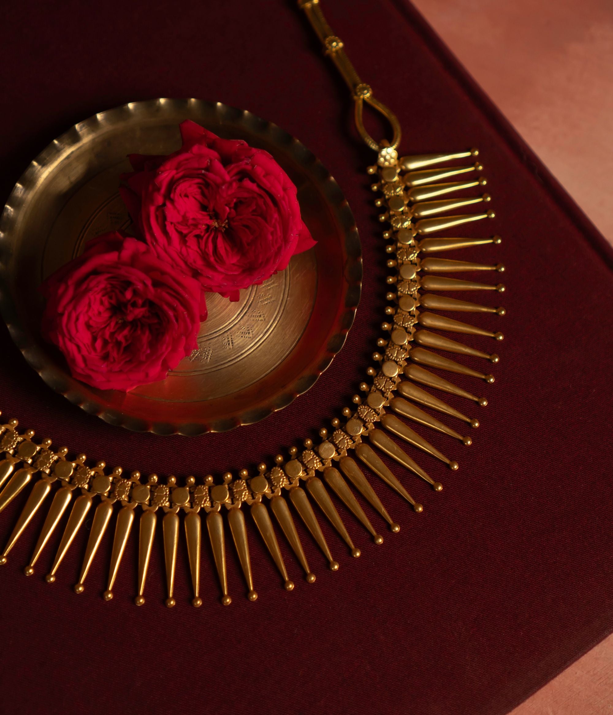 Gold Plated Silver Necklace with textured, spike-like pendants arranged in a semi-circle on a deep red surface. A brass dish and two red roses in the background enhance the rich, elegant aesthetic.