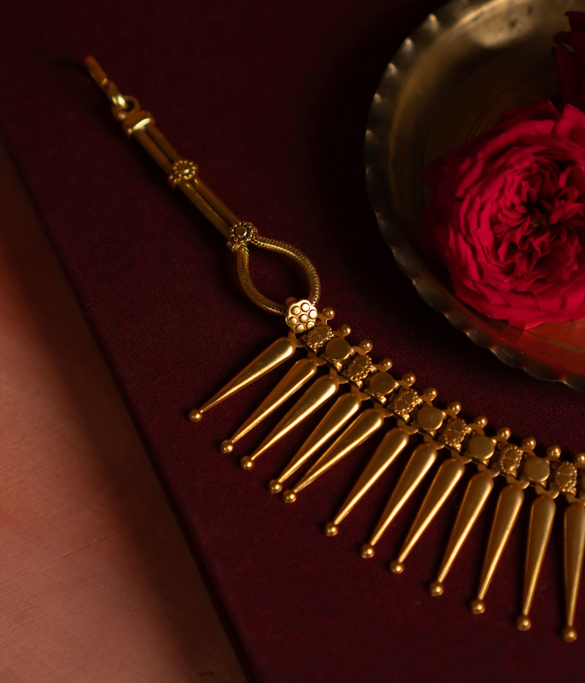 Gold Plated Silver Necklace with textured, spike-like pendants arranged in a semi-circle on a deep red surface. A brass dish and two red roses in the background enhance the rich, elegant aesthetic.