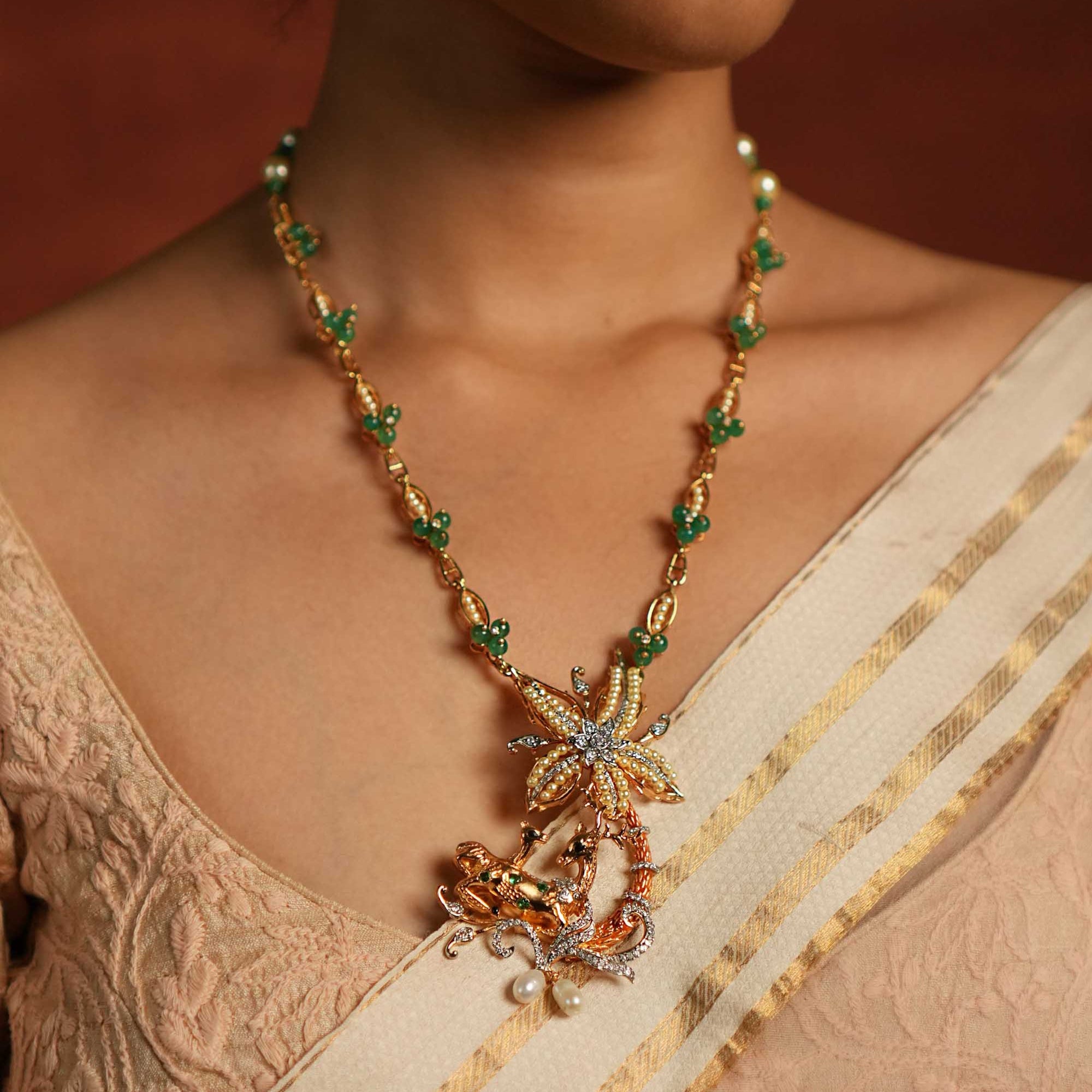 Model wearing a gold-toned floral pendant necklace with green stone accents, pearl detailing, and an ornate bird motif, styled with a beige saree.