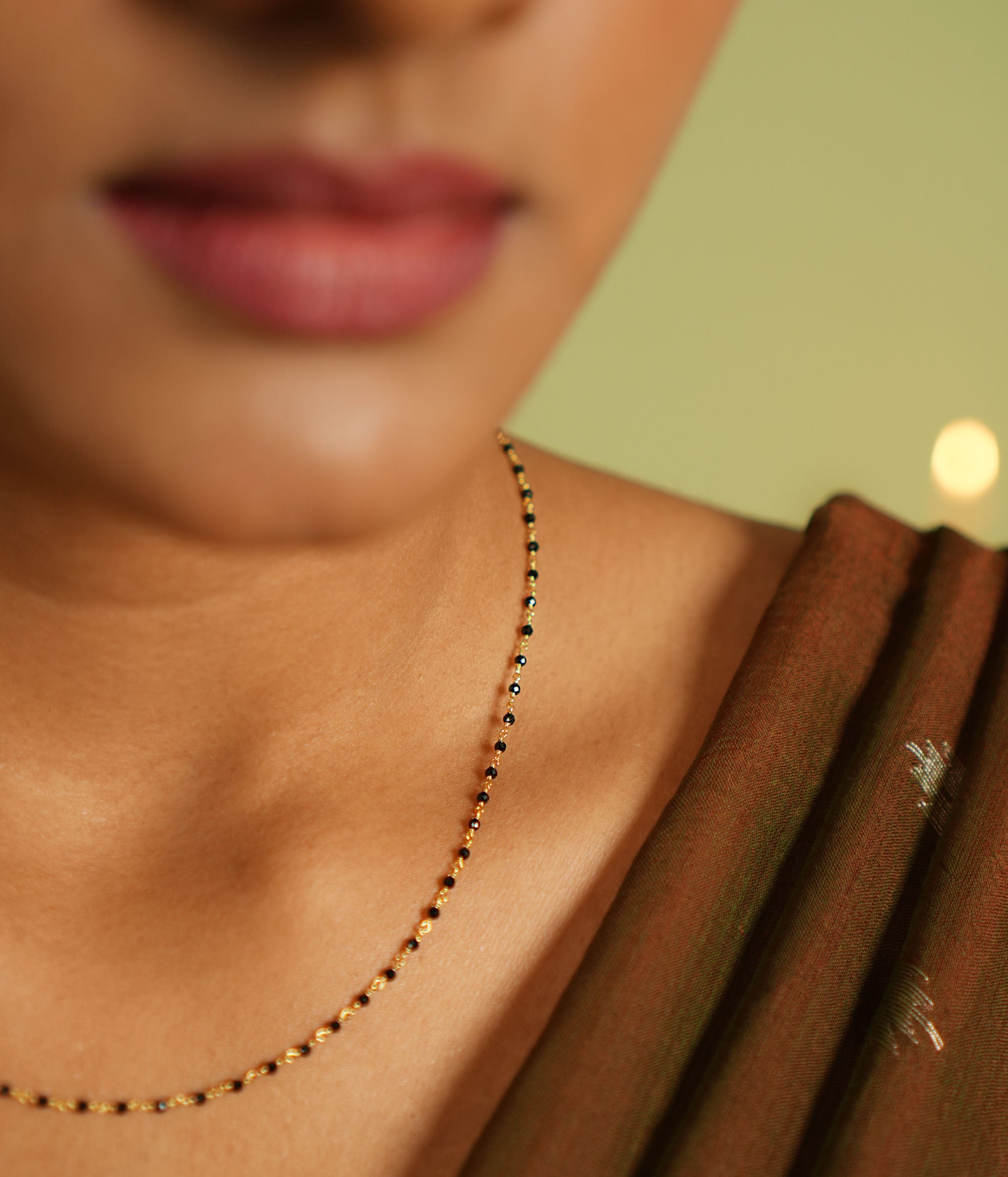 Woman wearing a delicate silver Mangal Sutra chain with gold plating, featuring black beads and subtle gold accents, resting gracefully on her neckline, paired with a muted saree in earthy tones.