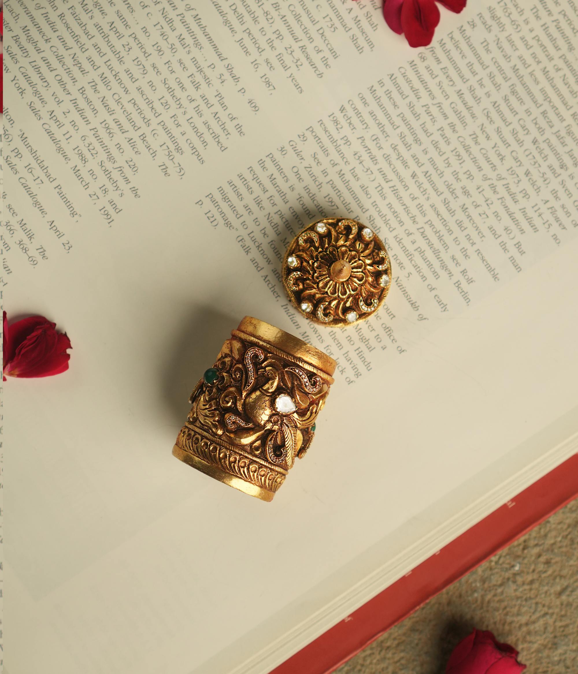 A meticulously crafted gold-plated silver sindoor case, adorned with floral polki and CZ stones on top, and peacock motifs with green kundan, radiates timeless elegance and cultural charm.