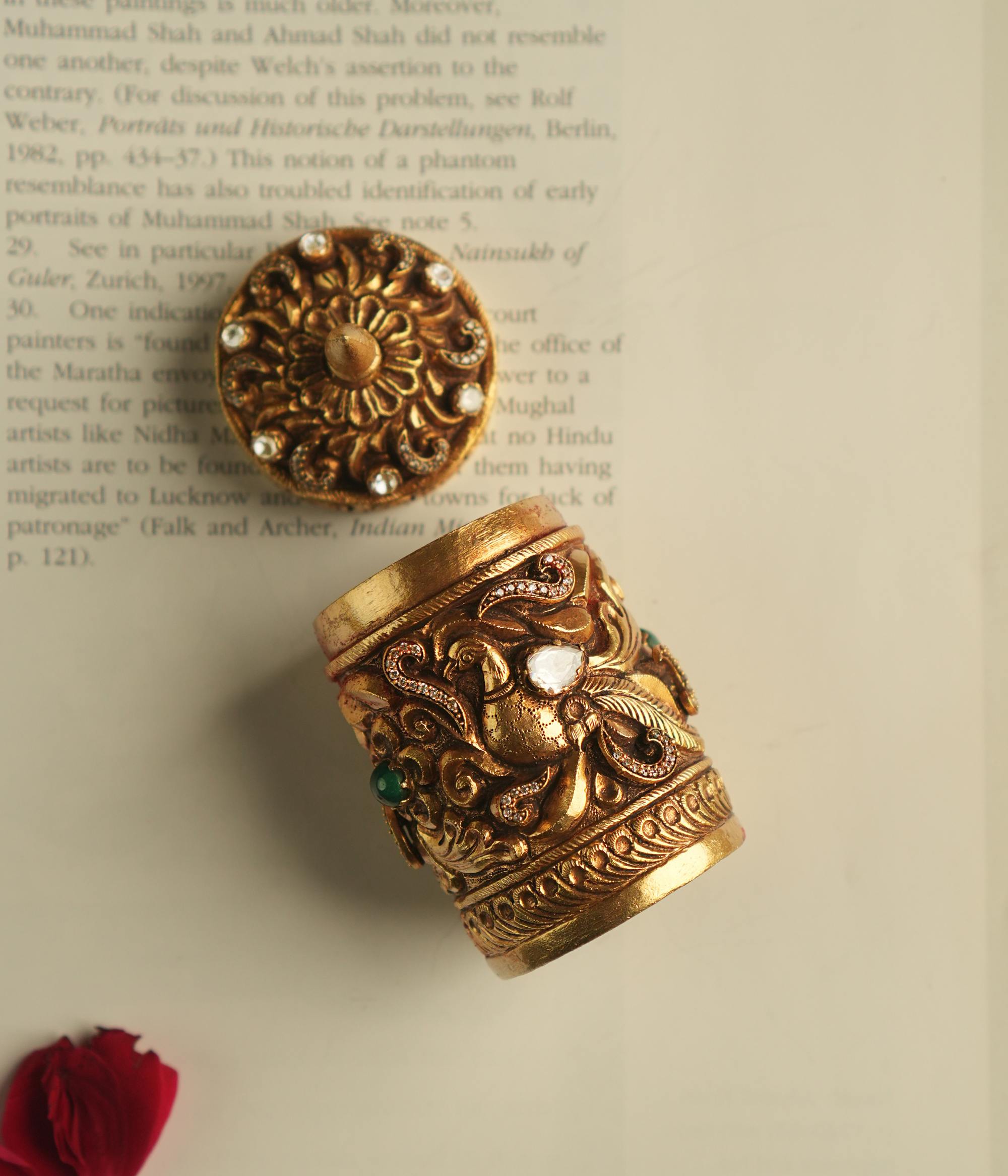 A meticulously crafted gold-plated silver sindoor case, adorned with floral polki and CZ stones on top, and peacock motifs with green kundan, radiates timeless elegance and cultural charm.