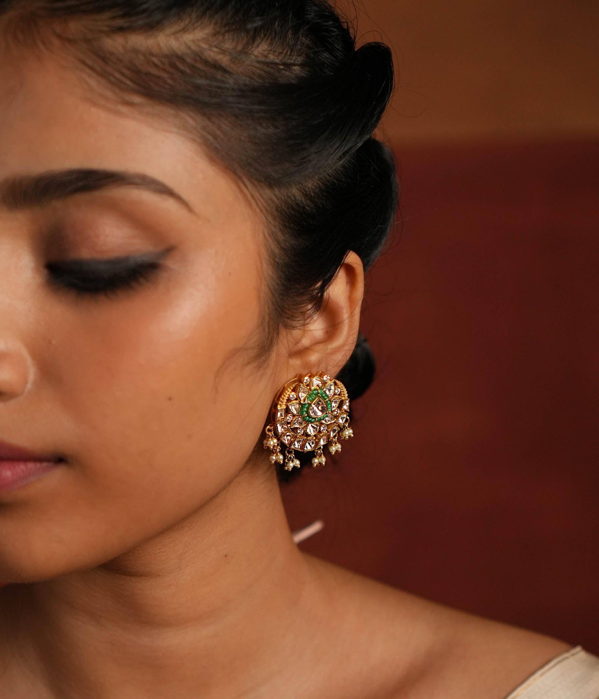 A beautifully crafted gold-plated silver drop earring featuring a dazzling Polki stone at the center, elegantly surrounded by a layer of shimmering CZ stones and vibrant green cut stones. The design is completed with delicate white pearl drops, adding a touch of grace and sophistication, making it a perfect accessory for both traditional and contemporary looks.