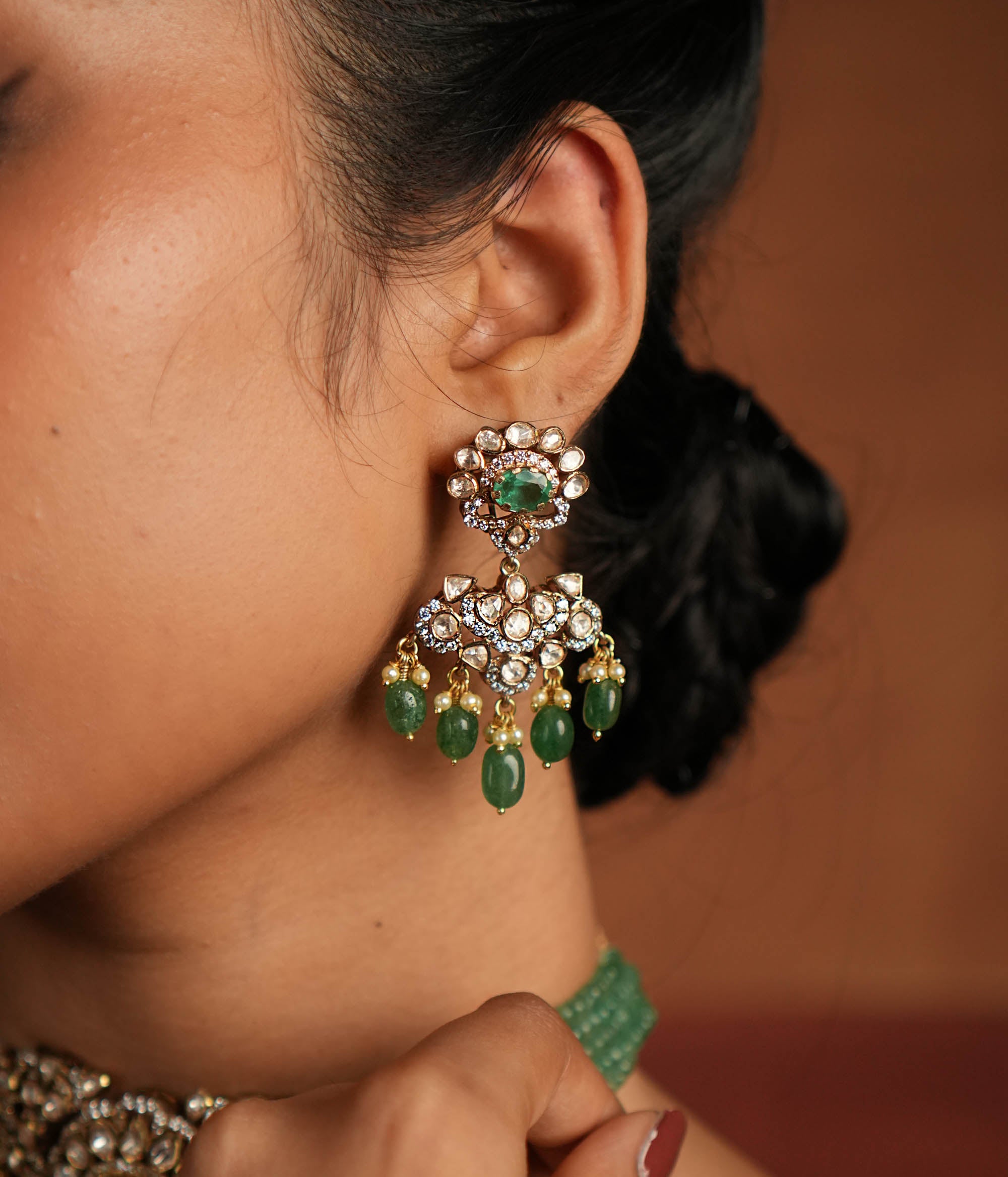 Victorian elegance reborn! These gold-plated silver dangler earrings feature a radiant green gemstone at the center, encircled by shimmering polki and CZ stones. Graceful pearl drops with golden beads enhance their charm, evoking the allure of a forgotten era. A timeless statement of beauty, crafted for the modern woman.