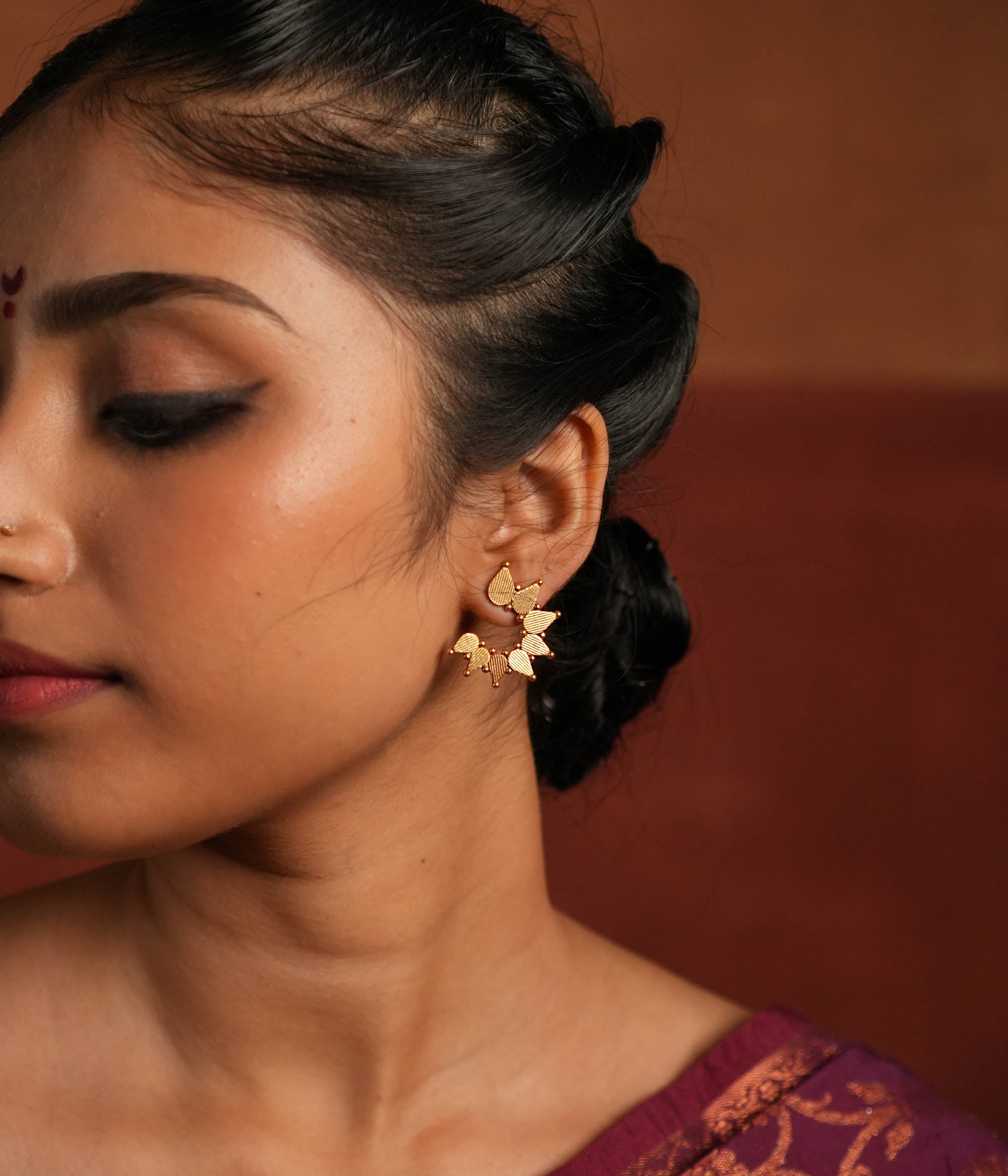 Gold-plated silver stud earrings featuring swirling leaf motifs intricately arranged in a circular dance, inspired by vintage Kolhapuri tribal designs. The detailed craftsmanship adds a touch of heritage charm, making them a timeless and elegant accessory.
