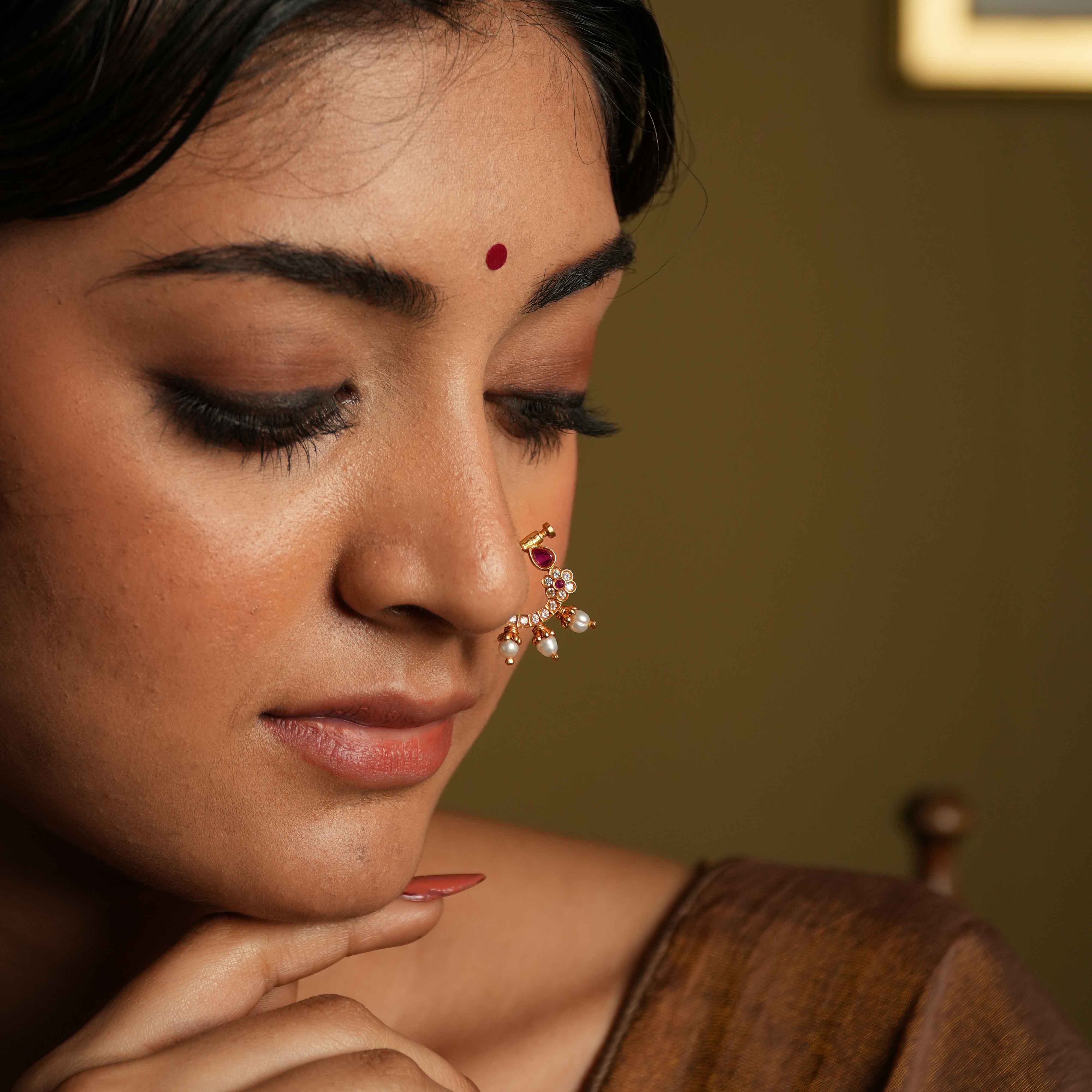 A gold-plated 92.5 sterling silver nose ring featuring a vintage South Indian-inspired design. It is adorned with CZ stones and red cut stones, set in a white Kundan setting. The piece includes a minimal flower motif with three bold pearl edgings, exuding timeless elegance and classic sophistication.