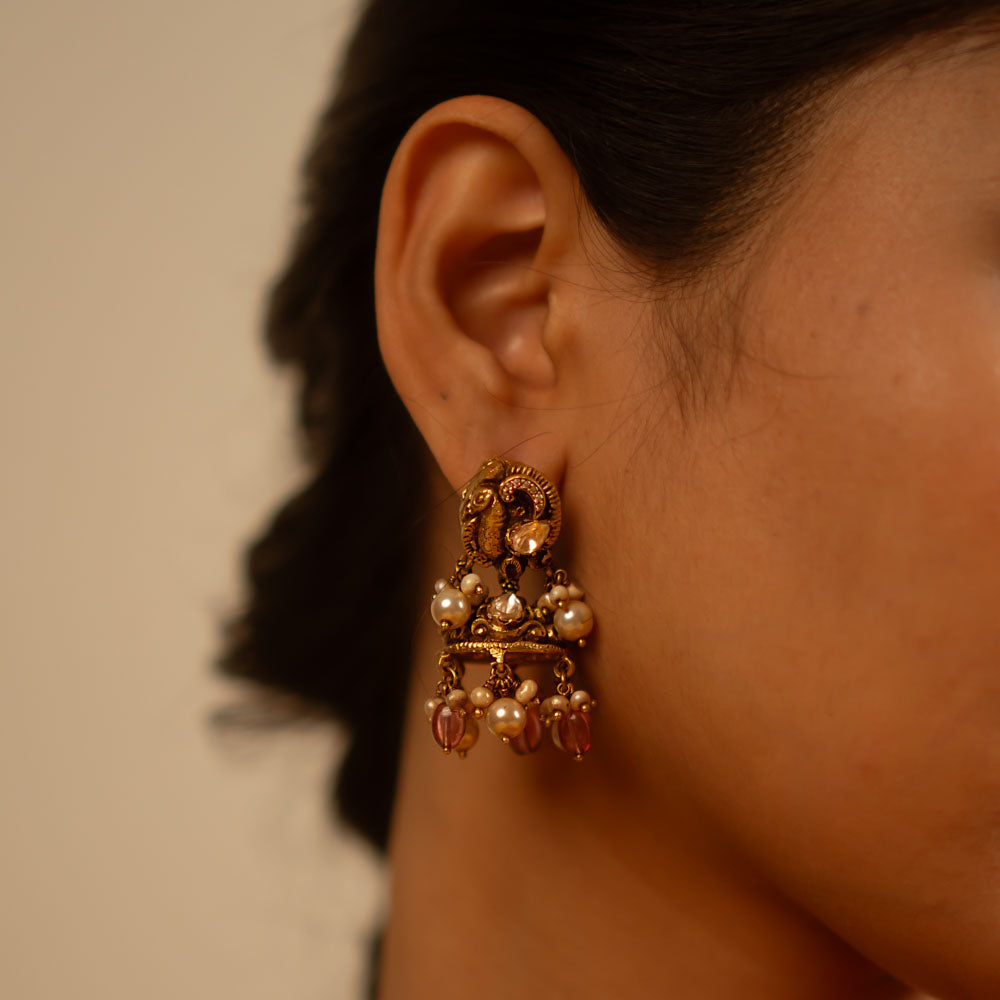 Handcrafted in antique gold-plated silver, these dangler earrings showcase a seamless fusion of classic elegance and modern charm. Adorned with white kundan stones, pearls, and red beads, their vintage-inspired design adds a timeless allure.