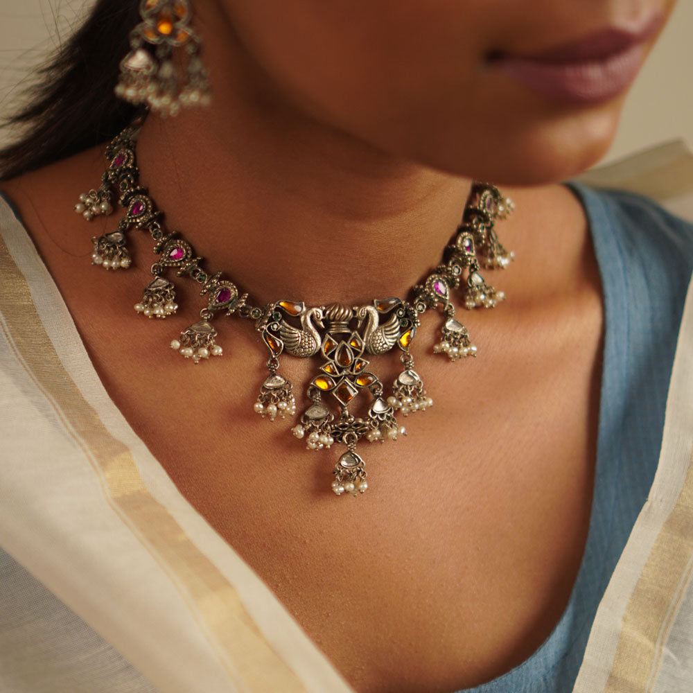 A cherished heirloom reimagined—the peacock-motif necklace in oxidized silver is adorned with radiant red and yellow kundan stones, finished with delicate pearl droplets. A timeless piece reminiscent of the treasured jewels passed down through generations.