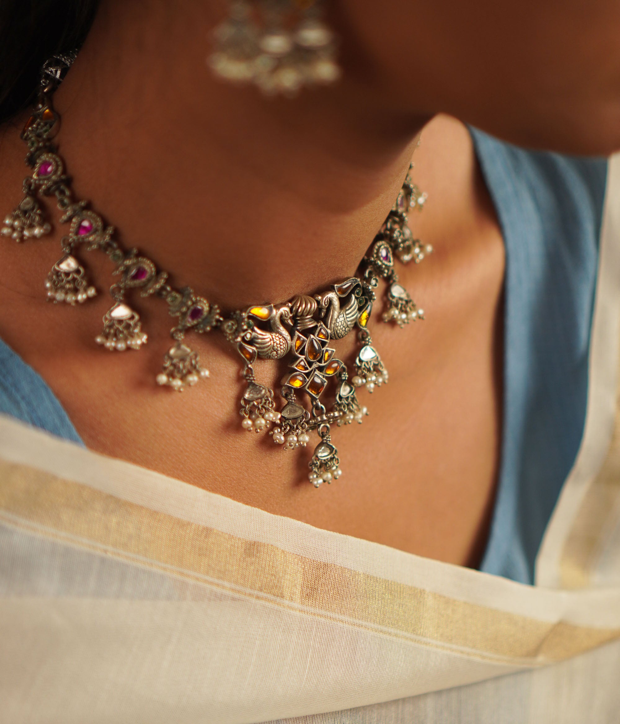 A cherished heirloom reimagined—the peacock-motif necklace in oxidized silver is adorned with radiant red and yellow kundan stones, finished with delicate pearl droplets. A timeless piece reminiscent of the treasured jewels passed down through generations.