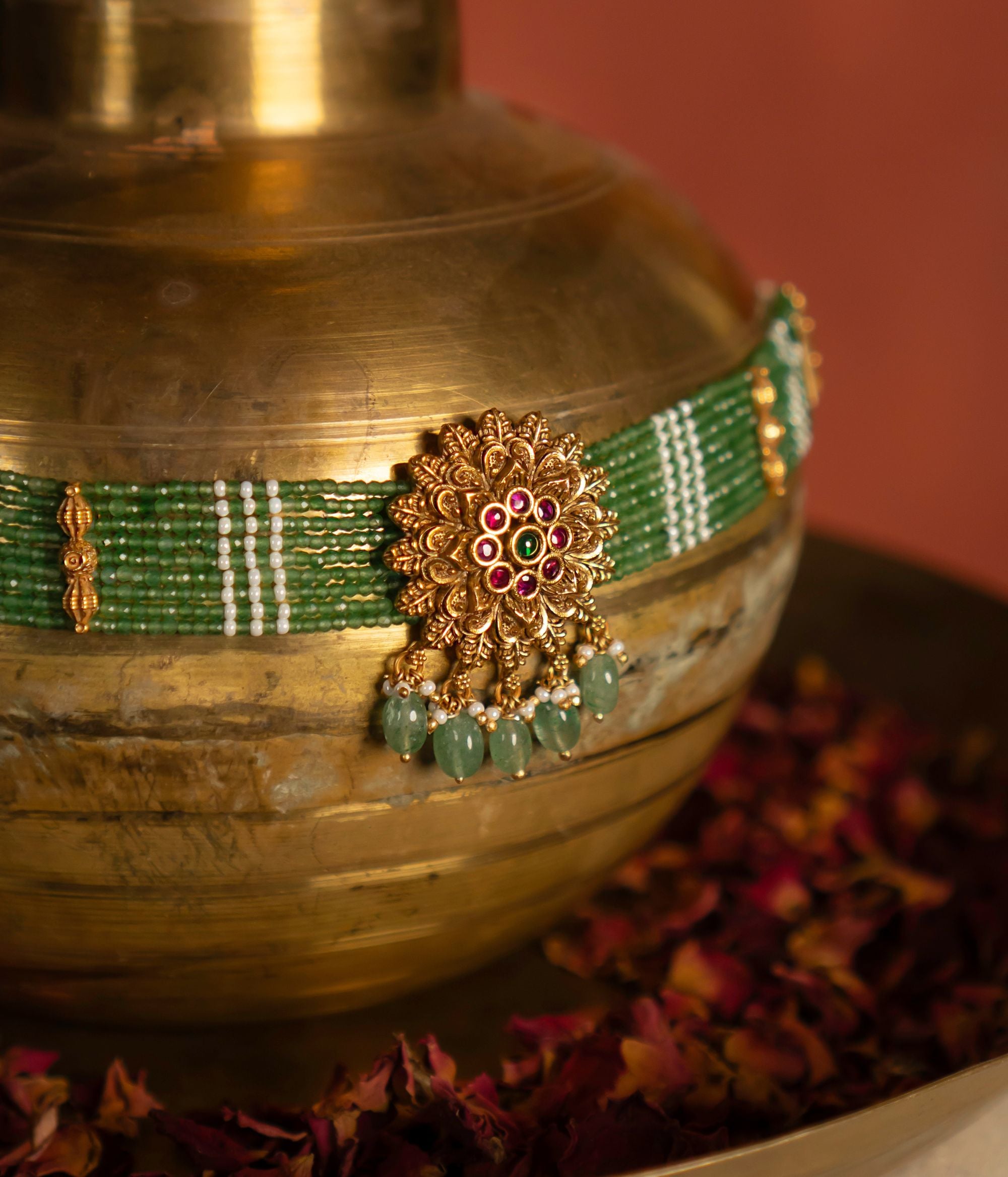 Gold-plated silver choker featuring a delicate green beaded chain with gold accents, leading to an ornate, round centerpiece adorned with red and green Kundan stones. The floral-inspired pendant is embellished with cascading drops of green quartz and pearls, adding a touch of elegance. Displayed on a rustic brass pot, with a hand subtly visible at the top, enhancing the artisanal charm.