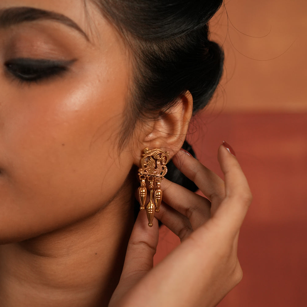 Tribal Majesty Reborn. These gold-plated silver dangler earrings, featuring a delicate peacock motif in the stud and a spike-like pendant with intricate designs, draw inspiration from vintage Kolhapuri artistry, blending timeless tradition with contemporary elegance.