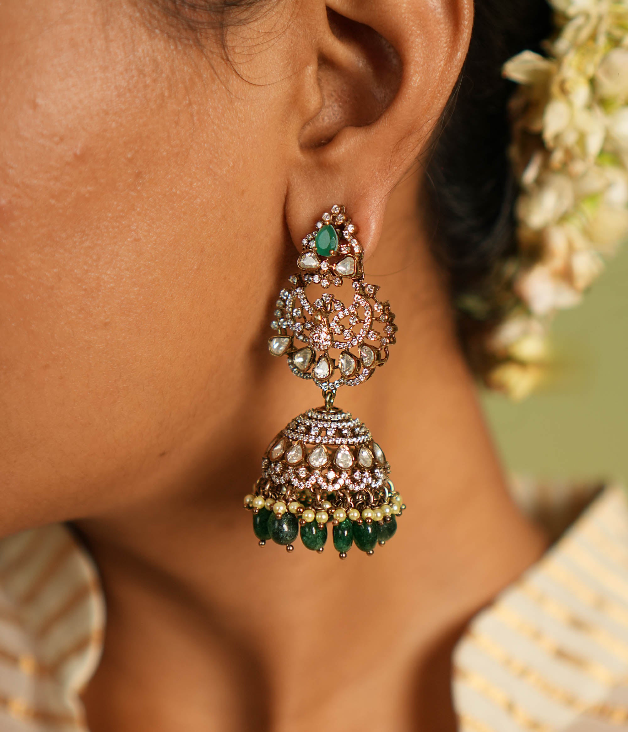 Exquisite gold-plated silver jhumkas featuring a graceful floral design, adorned with vibrant green gemstones, sparkling Polki stones, shimmering CZ stones, delicate pearls, and elegant green beads, offering a perfect blend of tradition and sophistication.