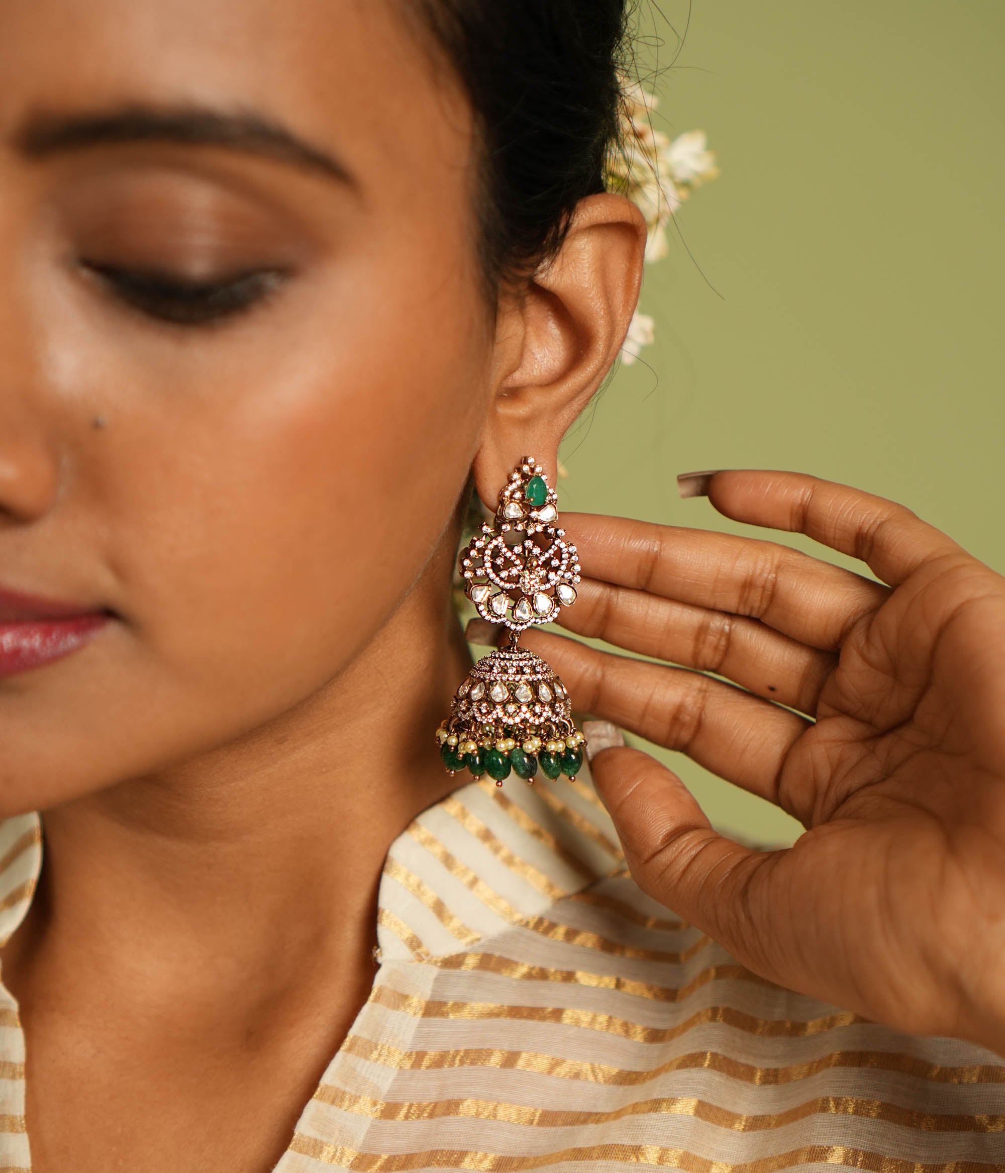 Exquisite gold-plated silver jhumkas featuring a graceful floral design, adorned with vibrant green gemstones, sparkling Polki stones, shimmering CZ stones, delicate pearls, and elegant green beads, offering a perfect blend of tradition and sophistication.