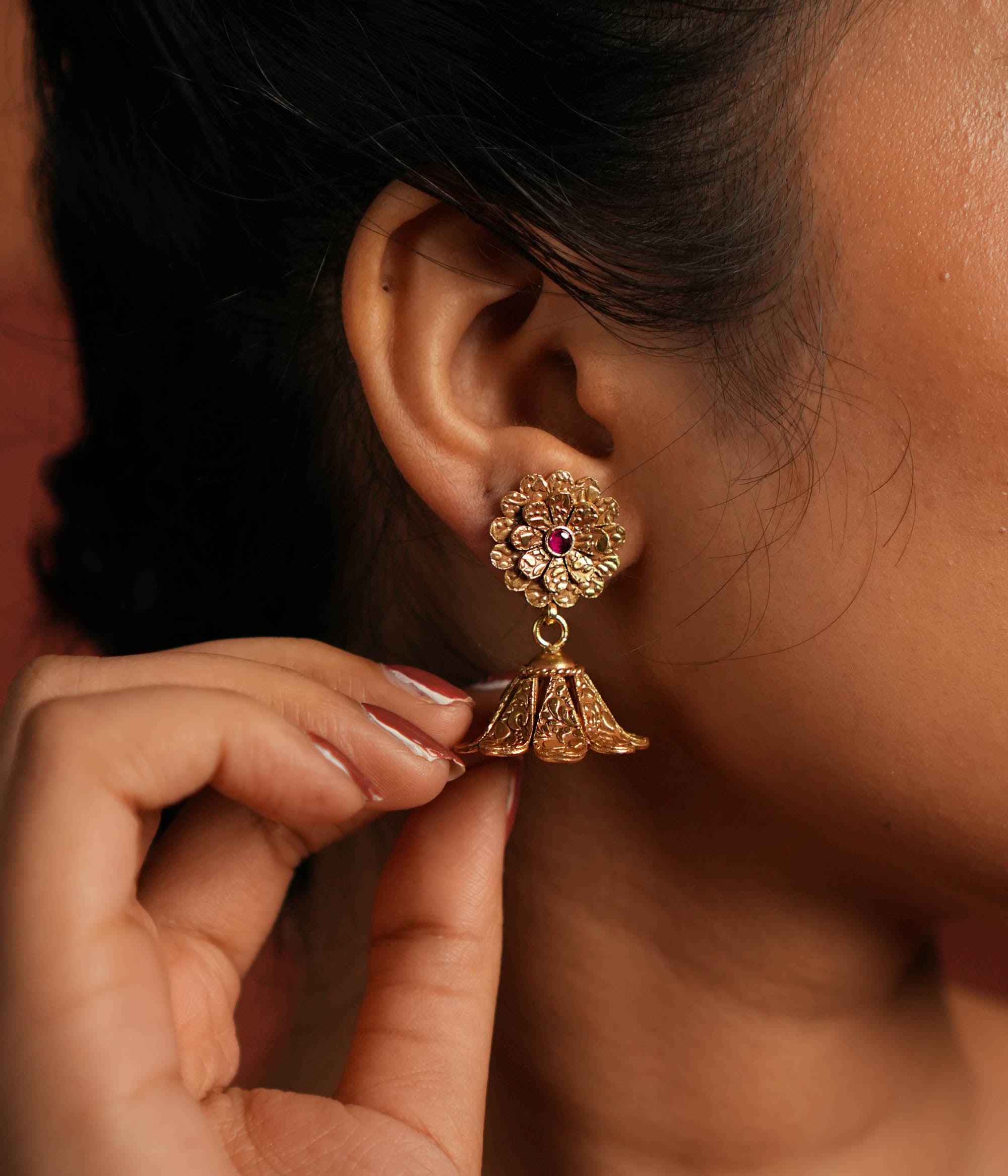 Exquisite jhumka earrings featuring a floral design engraved with intricate tiny floral details. The centerpiece showcases a striking red Kundan stone, enhancing the elegance of the design. The jhumka structure adds a traditional touch, making it a timeless statement piece.