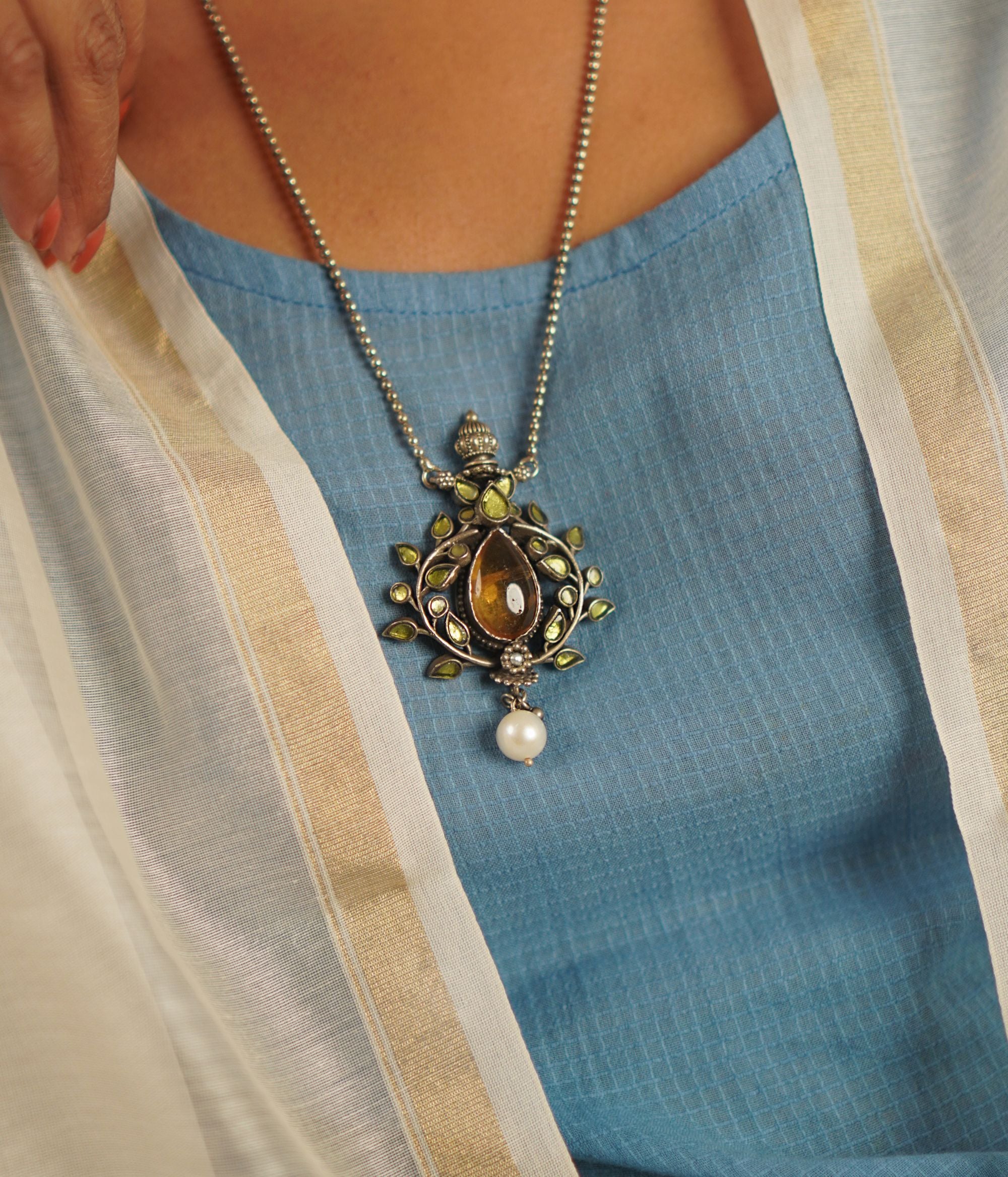This vintage-inspired silver pendant, suspended from a delicate beaded chain, features radiant yellow kundan, warm amber stones, and luminous pearls. Handcrafted with precision and care, it celebrates the charm of artisanal craftsmanship and the beauty of unique imperfections.