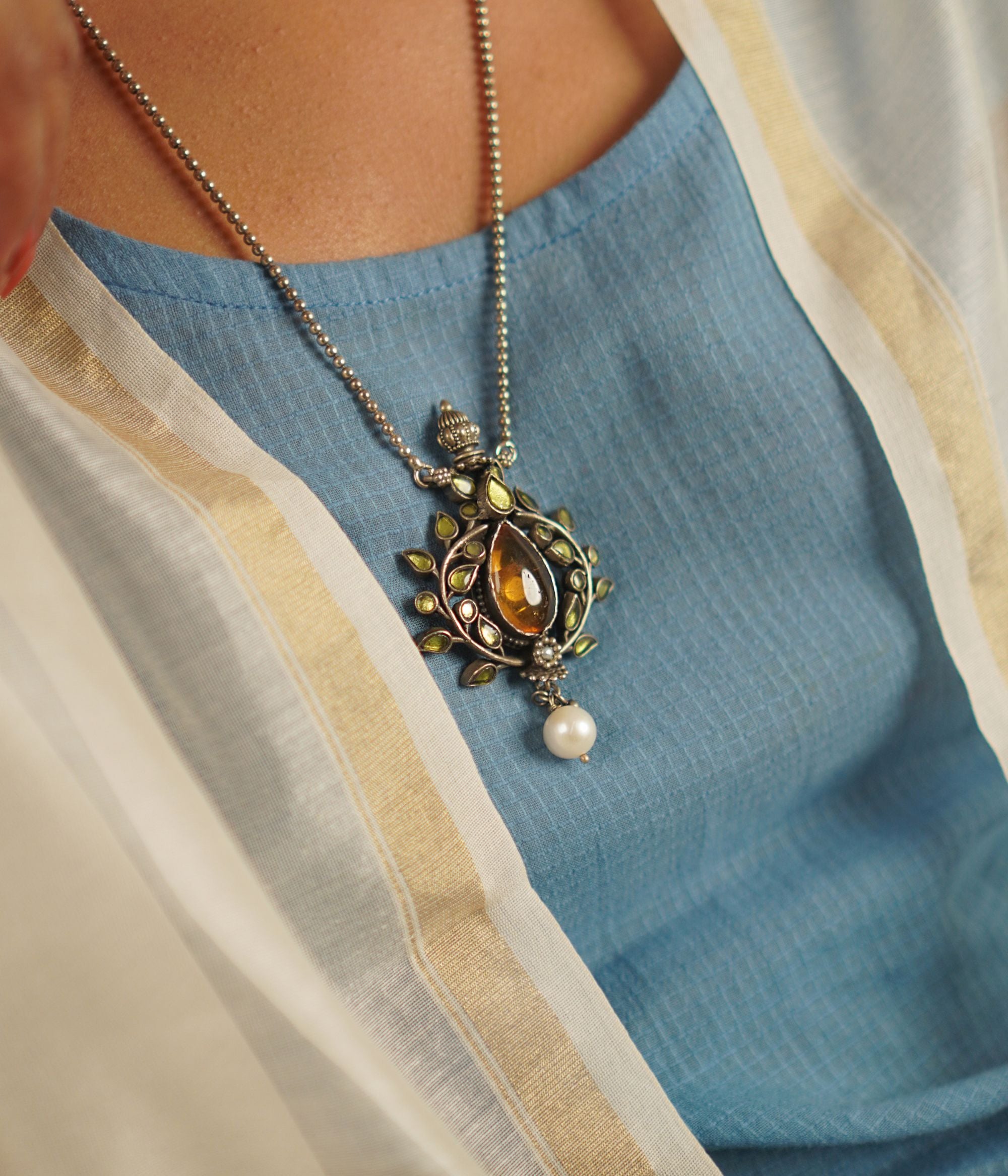 This vintage-inspired silver pendant, suspended from a delicate beaded chain, features radiant yellow kundan, warm amber stones, and luminous pearls. Handcrafted with precision and care, it celebrates the charm of artisanal craftsmanship and the beauty of unique imperfections.